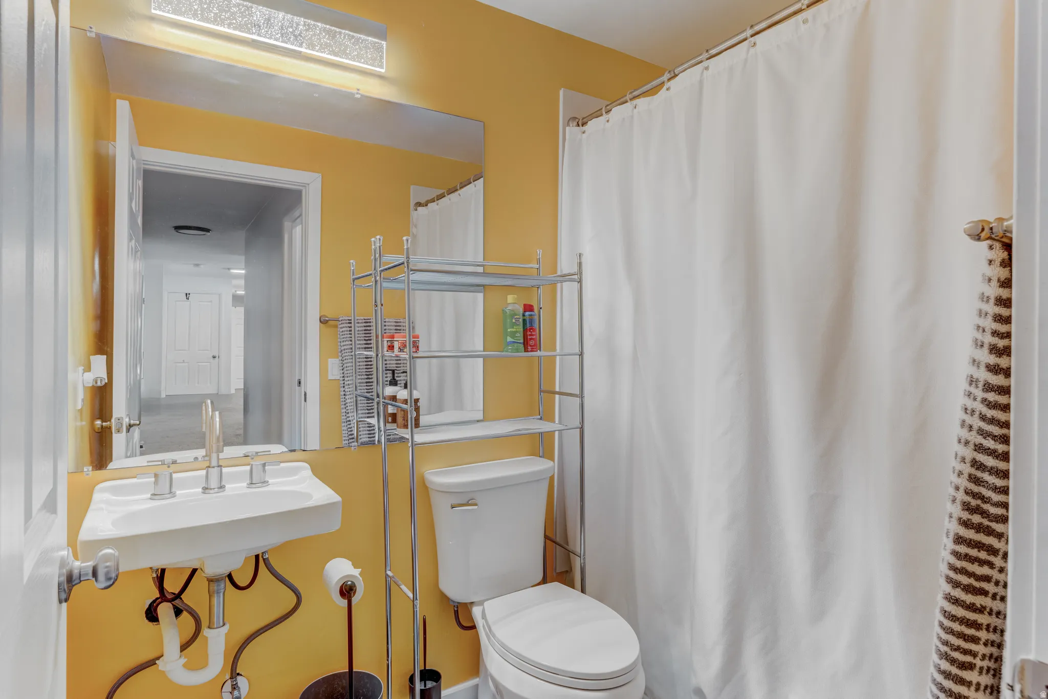 Bathroom featuring a wall-mounted sink, a toilet, a full-width mirror, a contemporary linear light fixture, and a shower curtain rod