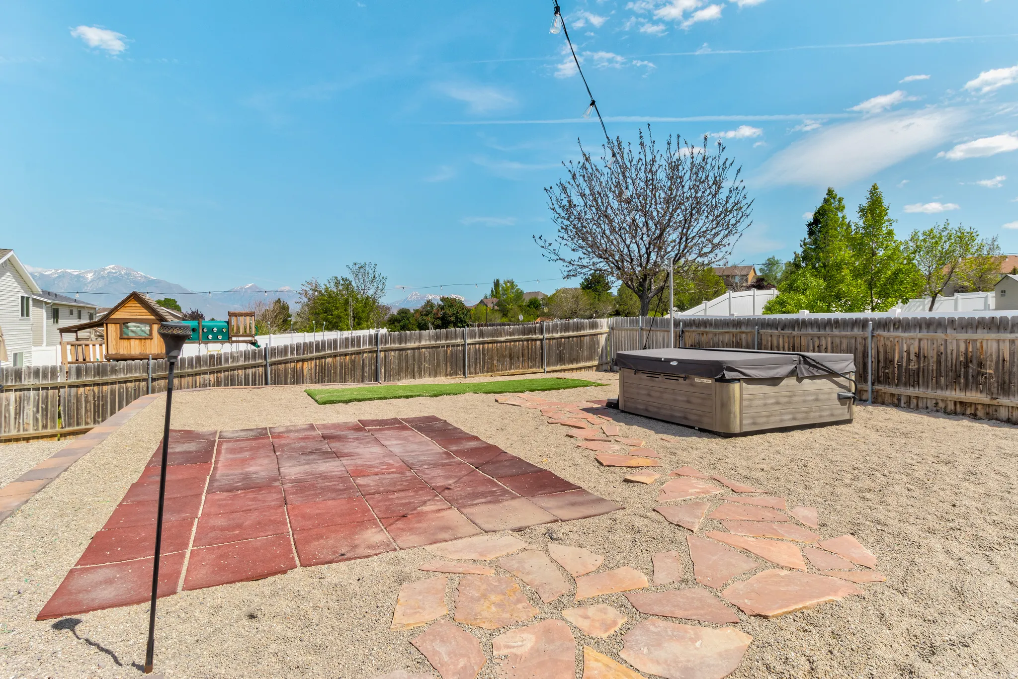 Expansive backyard featuring a hot tub, gravel ground cover, a natural wood privacy fence, stone paver pathways, and a mountain backdrop