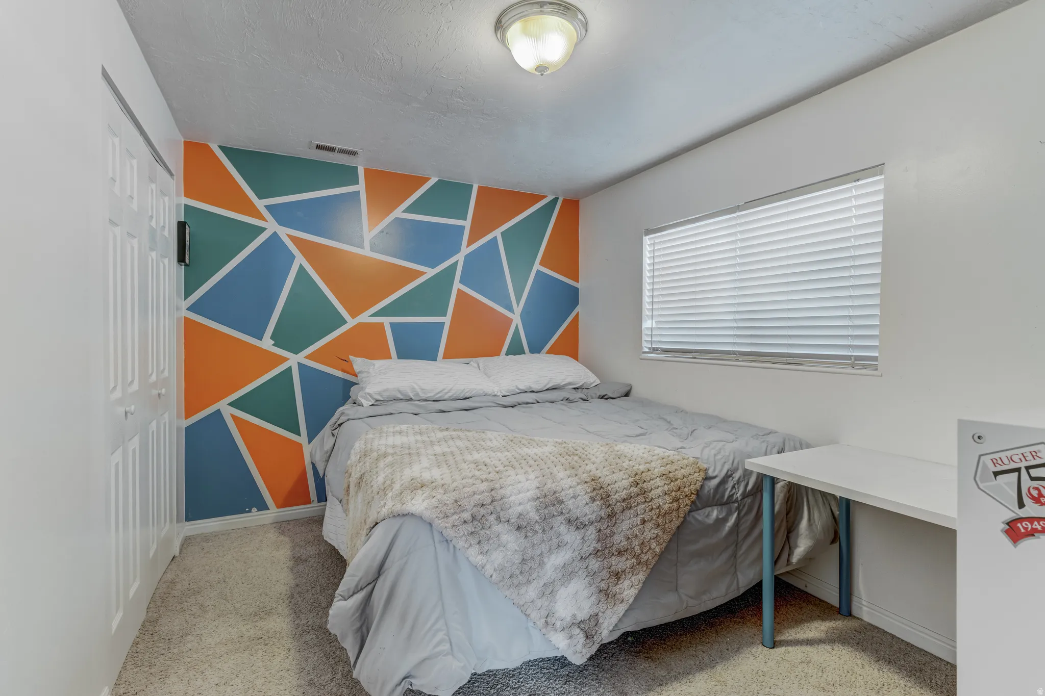 Bedroom featuring a geometric accent wall, bi-fold closet doors, wall-to-wall carpeting, and a single window with blinds