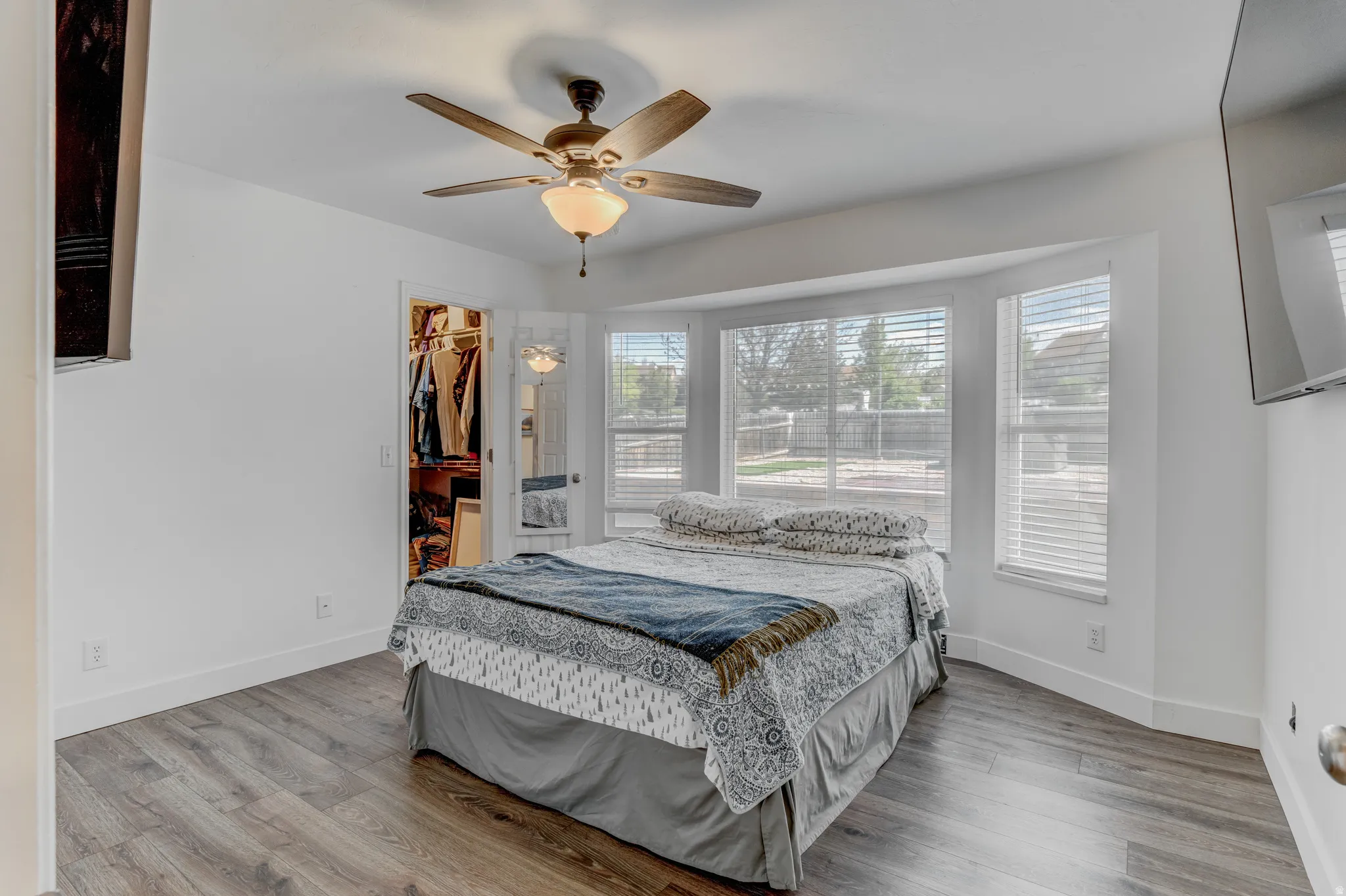 Bay window with multiple large windows, wood-finish flooring, ceiling fan with integrated lighting, recessed closet with bi-fold doors, and crisp white baseboards