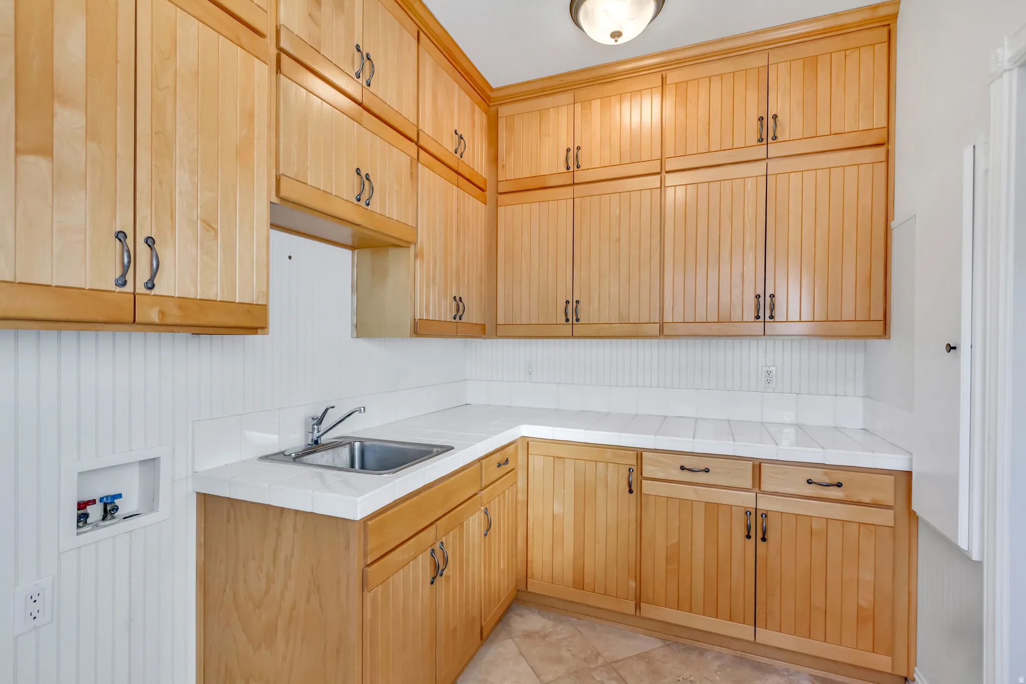 Utility room featuring extensive wood cabinetry, a stainless steel sink, tile countertops, beadboard wainscoting, and tile flooring
