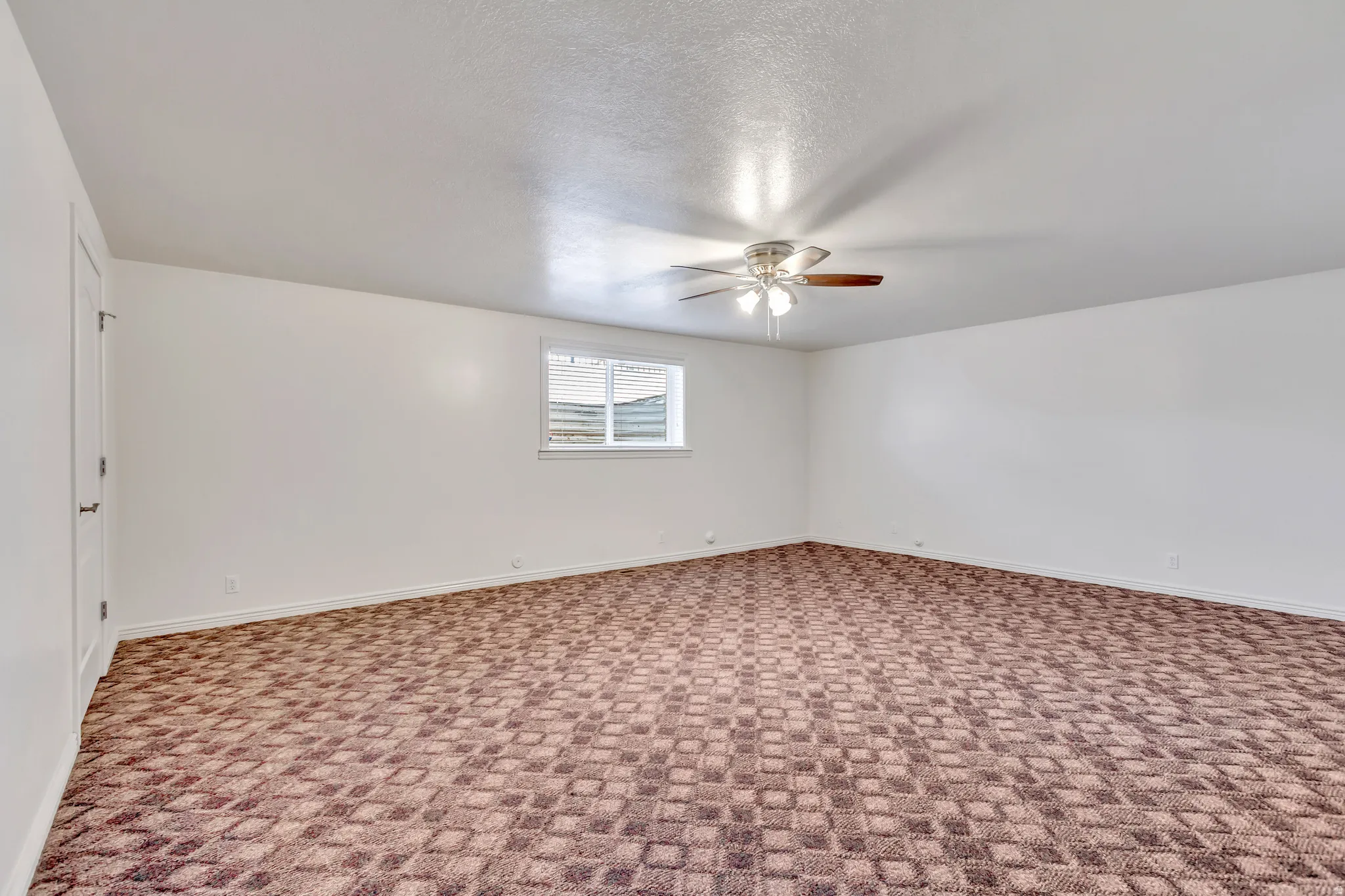 Spacious room featuring patterned carpet flooring, a window providing natural light, and a ceiling fan with integrated lighting