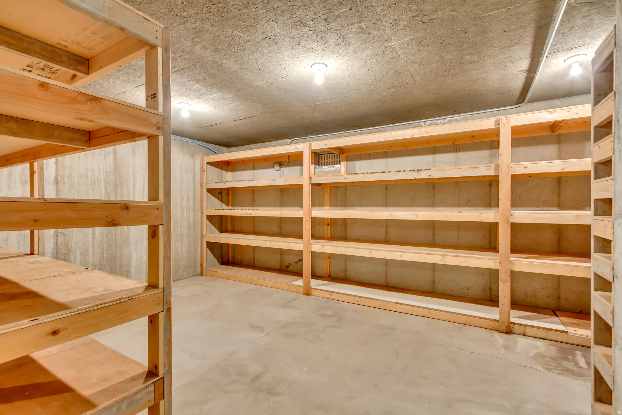 Basement storage area featuring extensive built-in wooden shelving, concrete walls, concrete flooring, and ceiling-mounted lighting fixtures