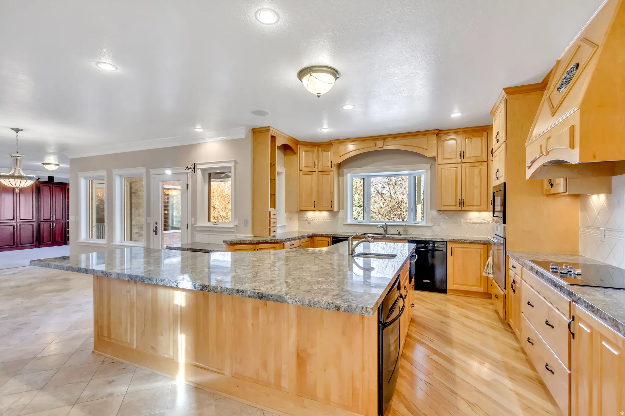Expansive kitchen featuring light wood cabinetry, stone countertops, integrated appliances, a built-in cooktop, and contrasting tile and wood-finish flooring