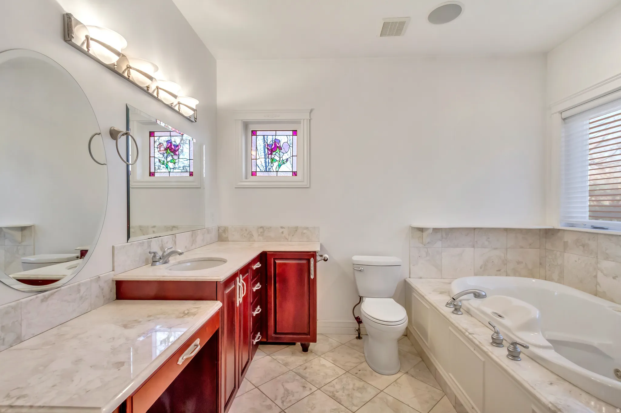 Full bathroom featuring a single vanity with wood-finish cabinetry, a built-in jetted tub, tiled flooring, and decorative stained-glass windows