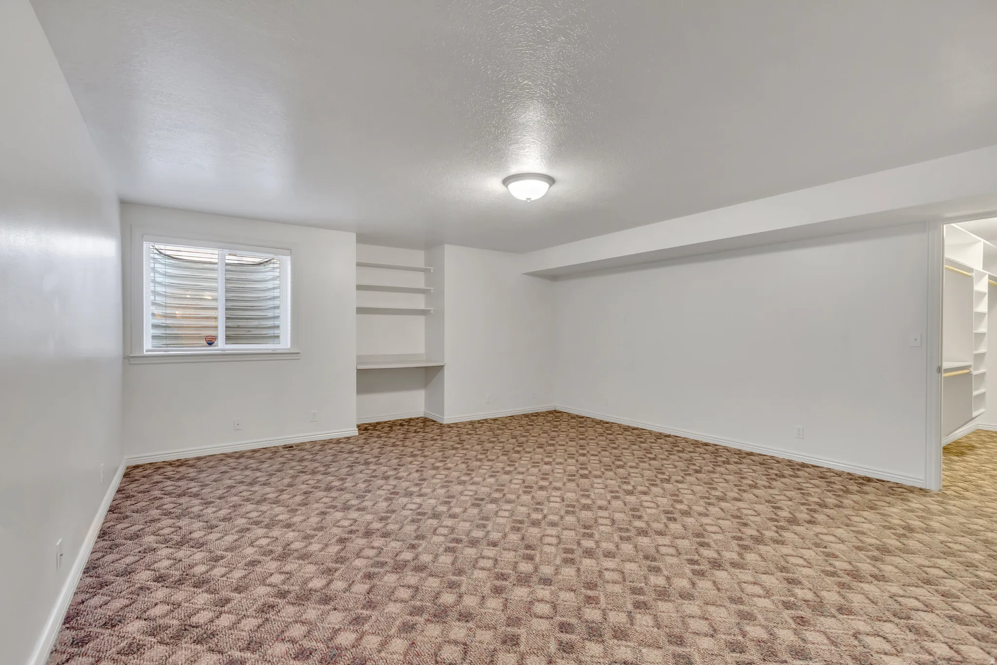 Spacious room featuring patterned carpeting, recessed ceiling light fixture, a window with horizontal blinds, built-in shelving, and a built-in closet system