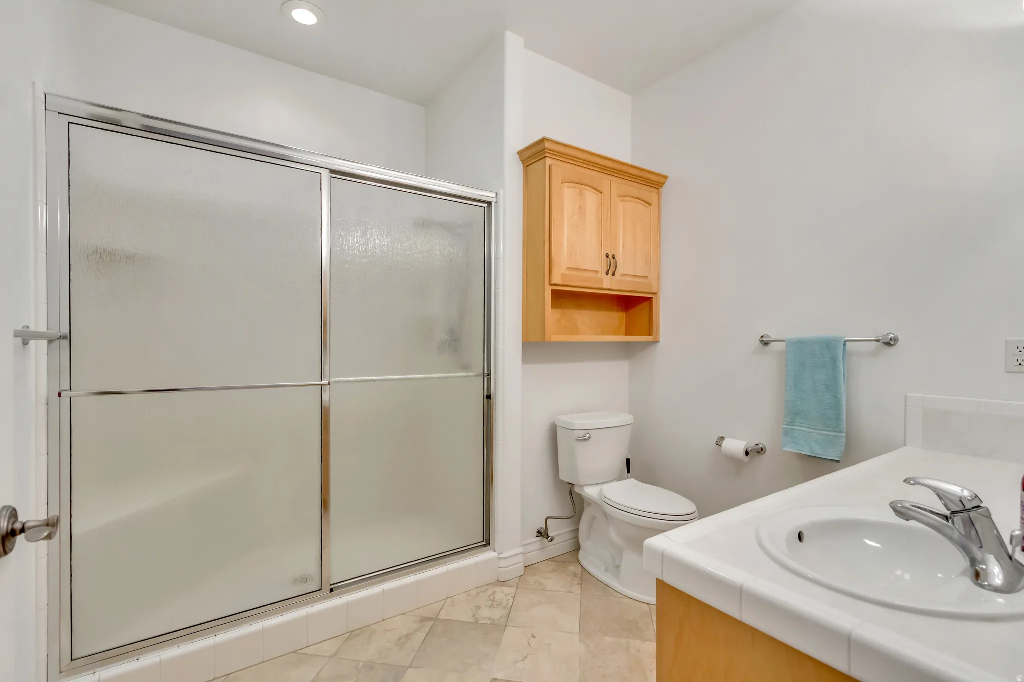 Bathroom featuring a walk-in shower with sliding frosted glass doors, light wood cabinetry, a white countertop sink, and tile flooring