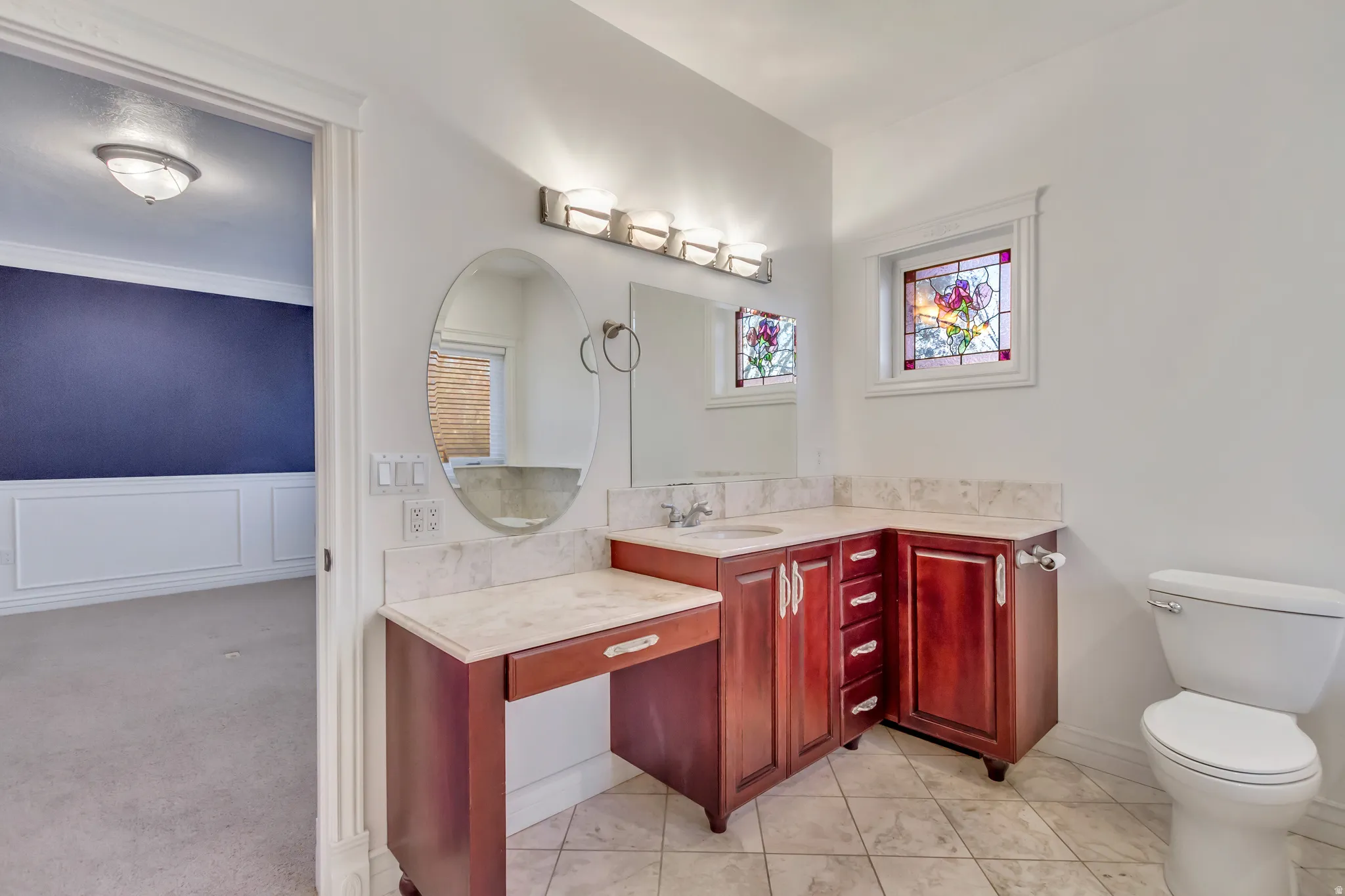 Bathroom featuring a vanity with a white countertop, rich wood-tone cabinetry, tiled flooring, a stained-glass window, and a wall-mounted light fixture
