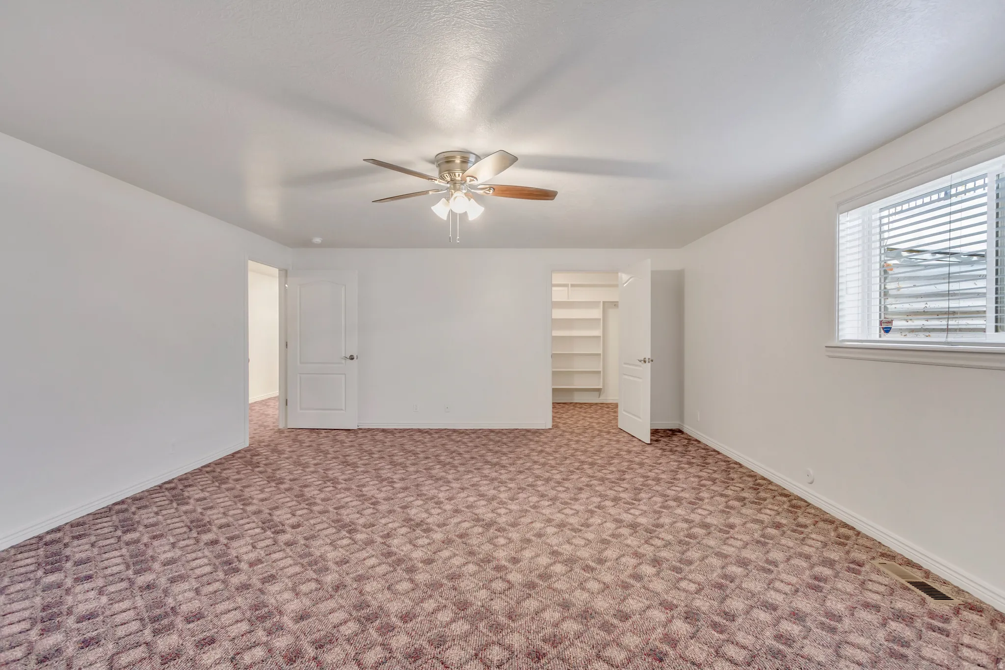 Spacious room featuring patterned carpet flooring, a ceiling fan with light fixture, and a window with horizontal blinds