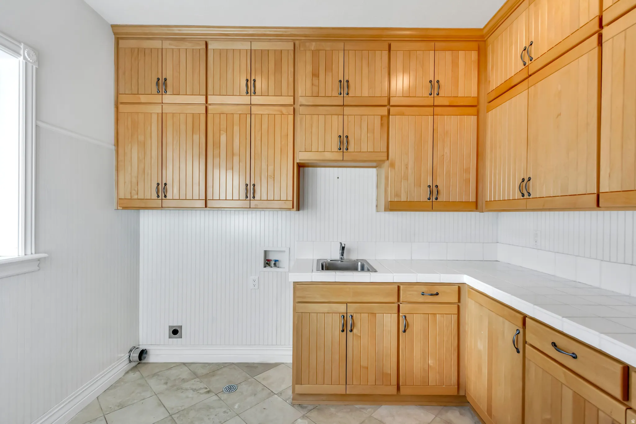 Utility room featuring extensive natural wood cabinetry, white beadboard paneling, a stainless steel utility sink, white countertops, and light-toned floor tiling