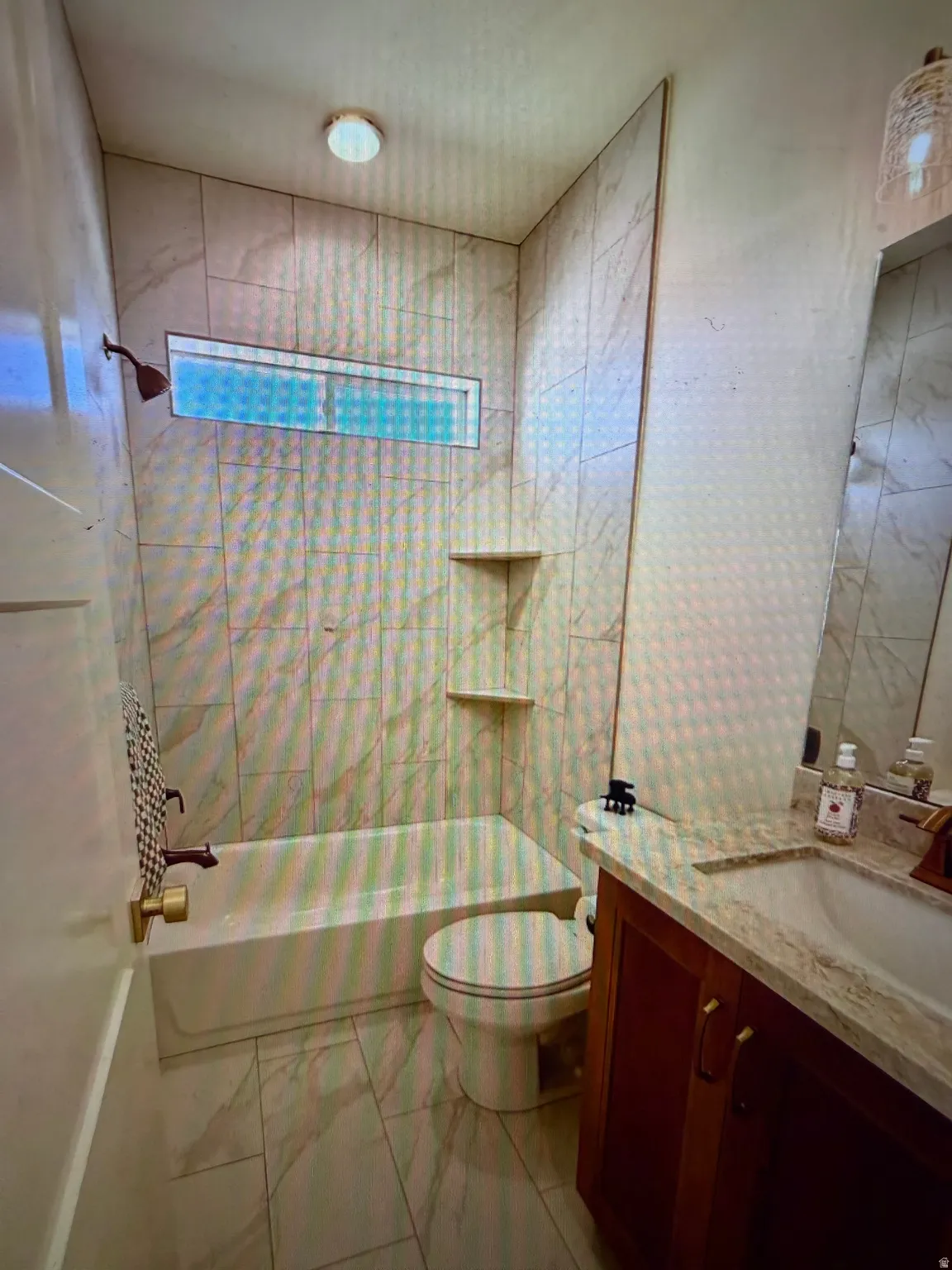 Bathroom featuring a combination shower/tub with marble-look tile surround, corner shelving, and a horizontal privacy window