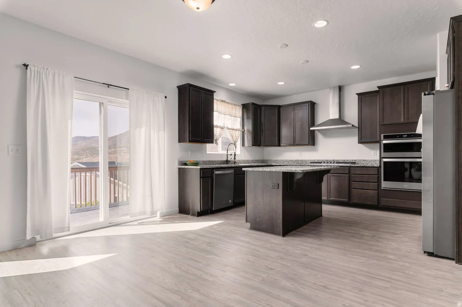 Open-concept kitchen featuring wood-finish flooring, dark wood cabinetry, a central island with granite-style countertops, stainless steel appliances, and recessed lighting