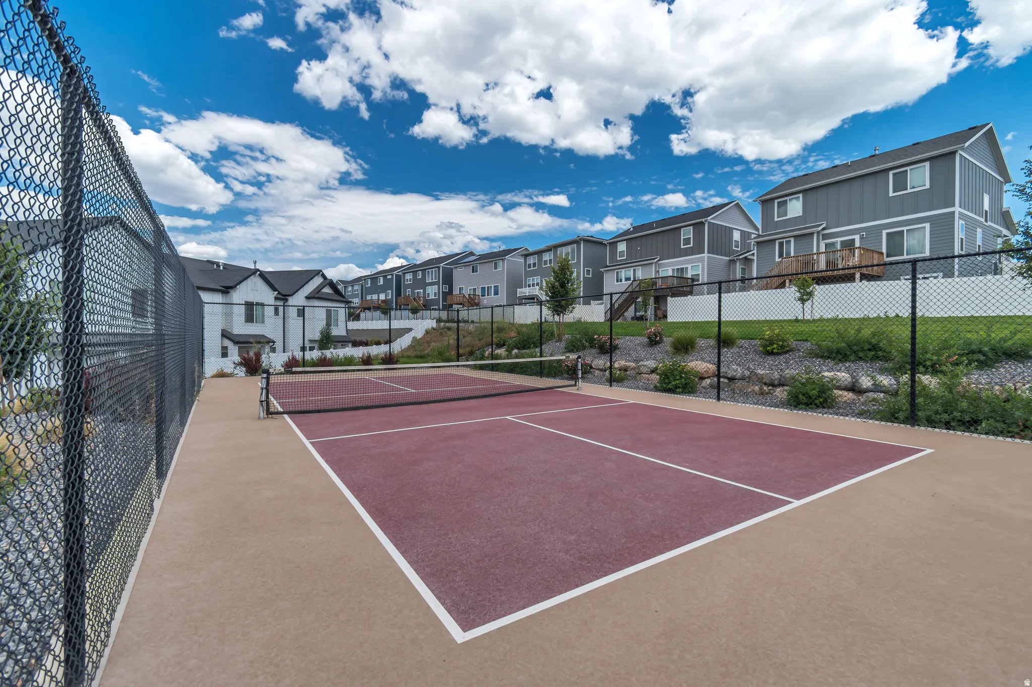 Community pickleball court featuring a red playing surface with white boundary lines, surrounded by a black chain-link fence