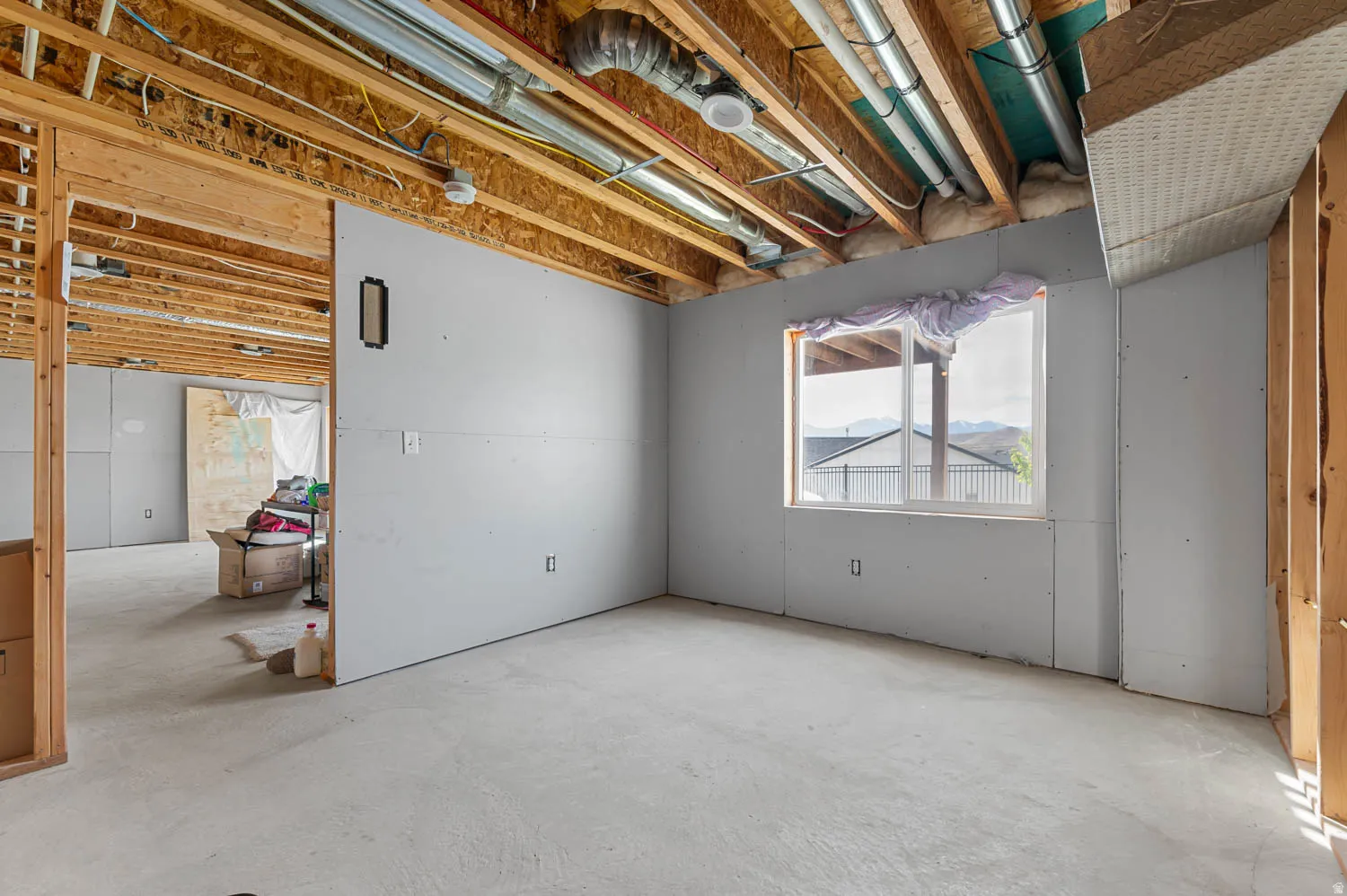 Unfinished lower level featuring exposed wood joists, concrete flooring, partially installed drywall, and a window offering a distant landscape view