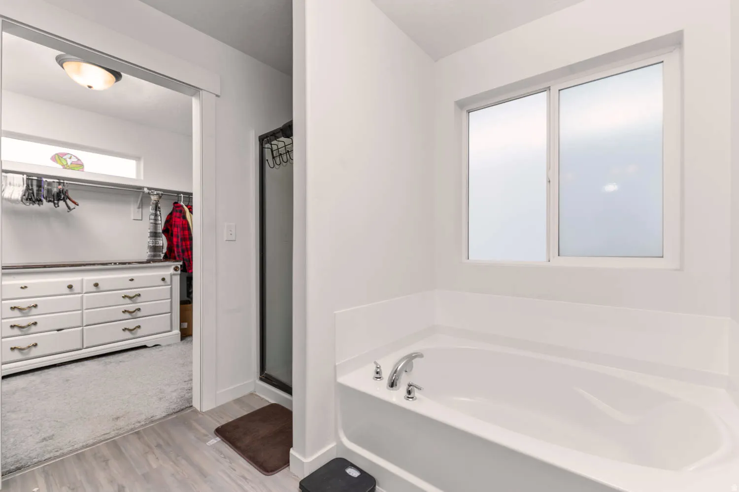 Soaking tub with chrome fixtures, frosted glass window, and wood-finish flooring