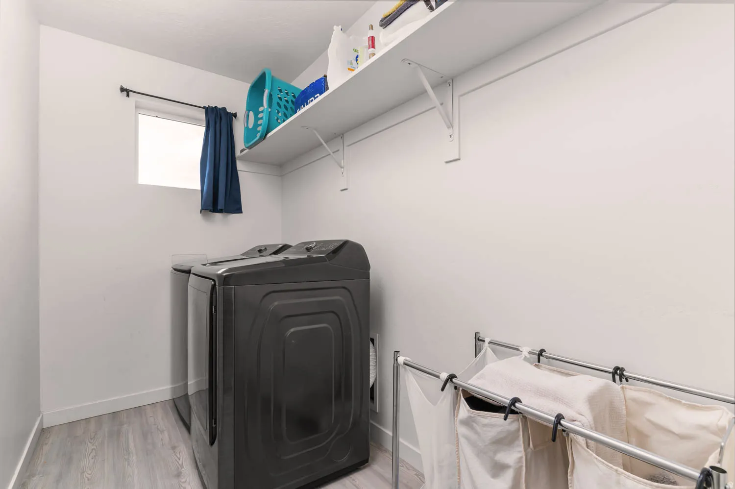 Dedicated laundry area featuring light-toned wood-finish flooring, a single window, white walls, and an overhead storage shelf