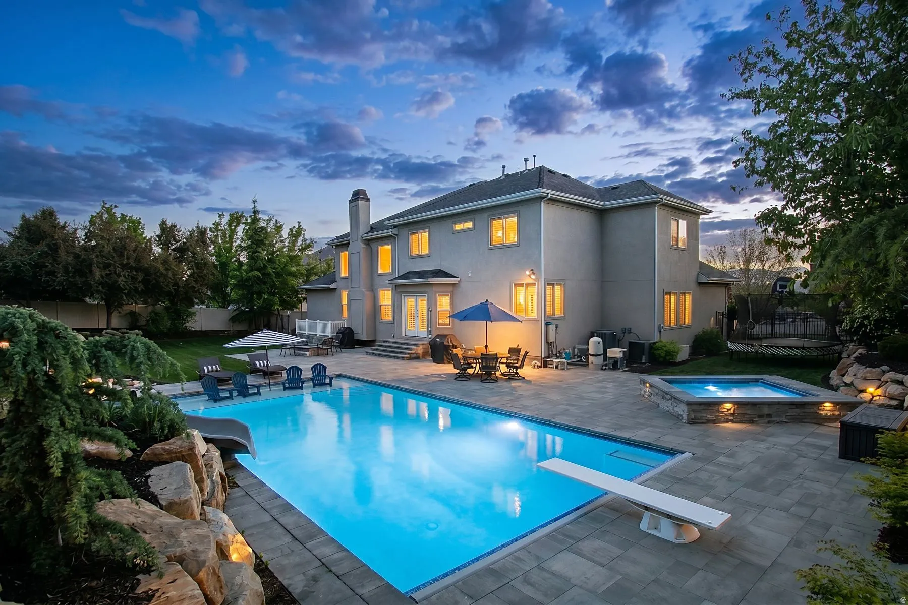 Expansive paved patio featuring a large swimming pool with a diving board, an integrated spa, and mature landscaping