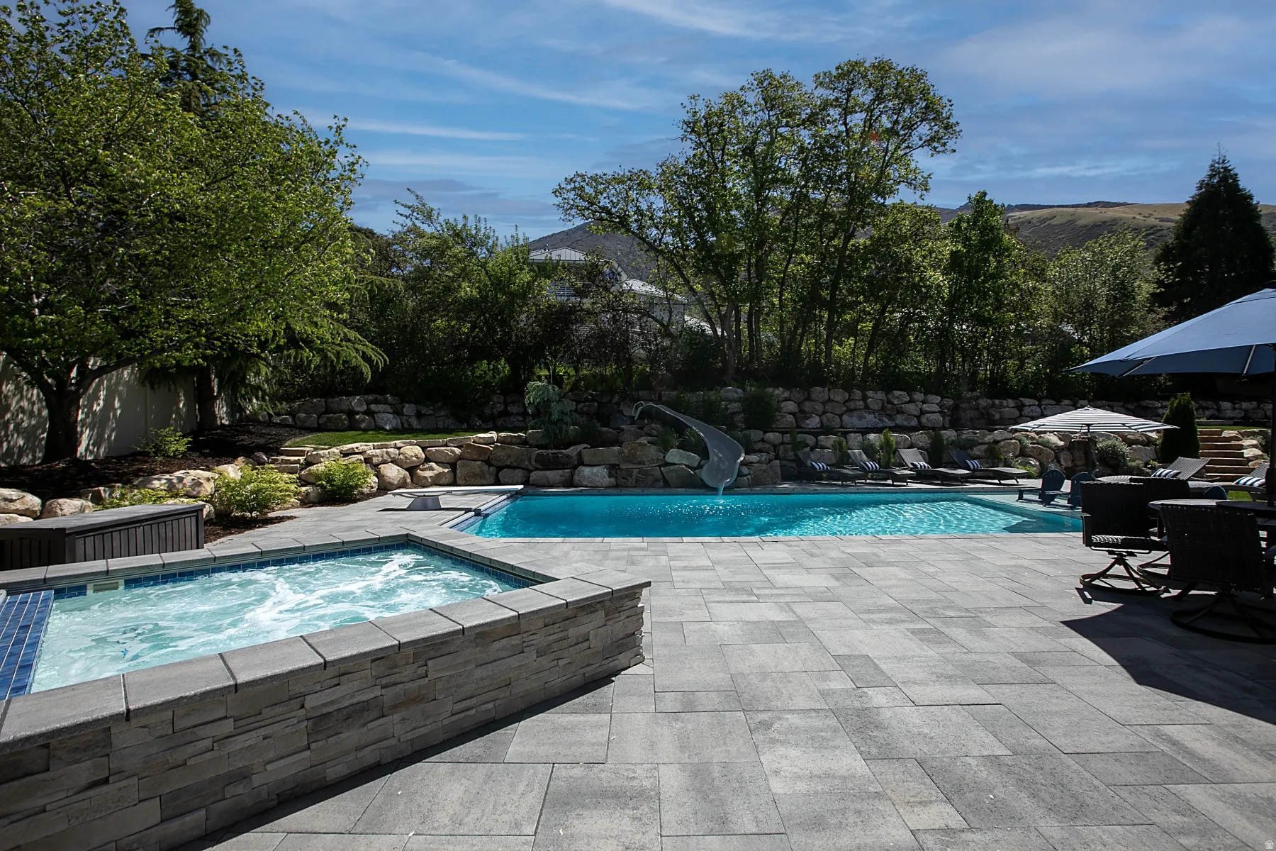 Outdoor living area featuring a rectangular pool, integrated spa with bubbling water, and a rock-lined water slide