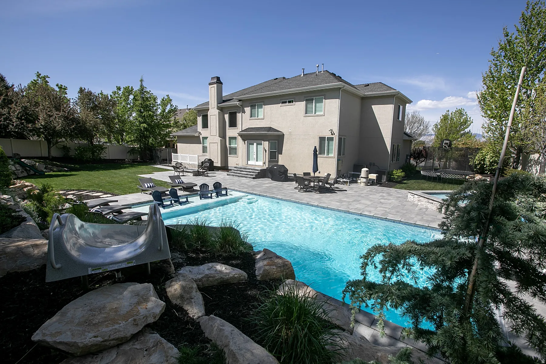 Expansive backyard featuring a large swimming pool with integrated spa, extensive paver patio surrounds, a substantial lawn area, and mature landscaping