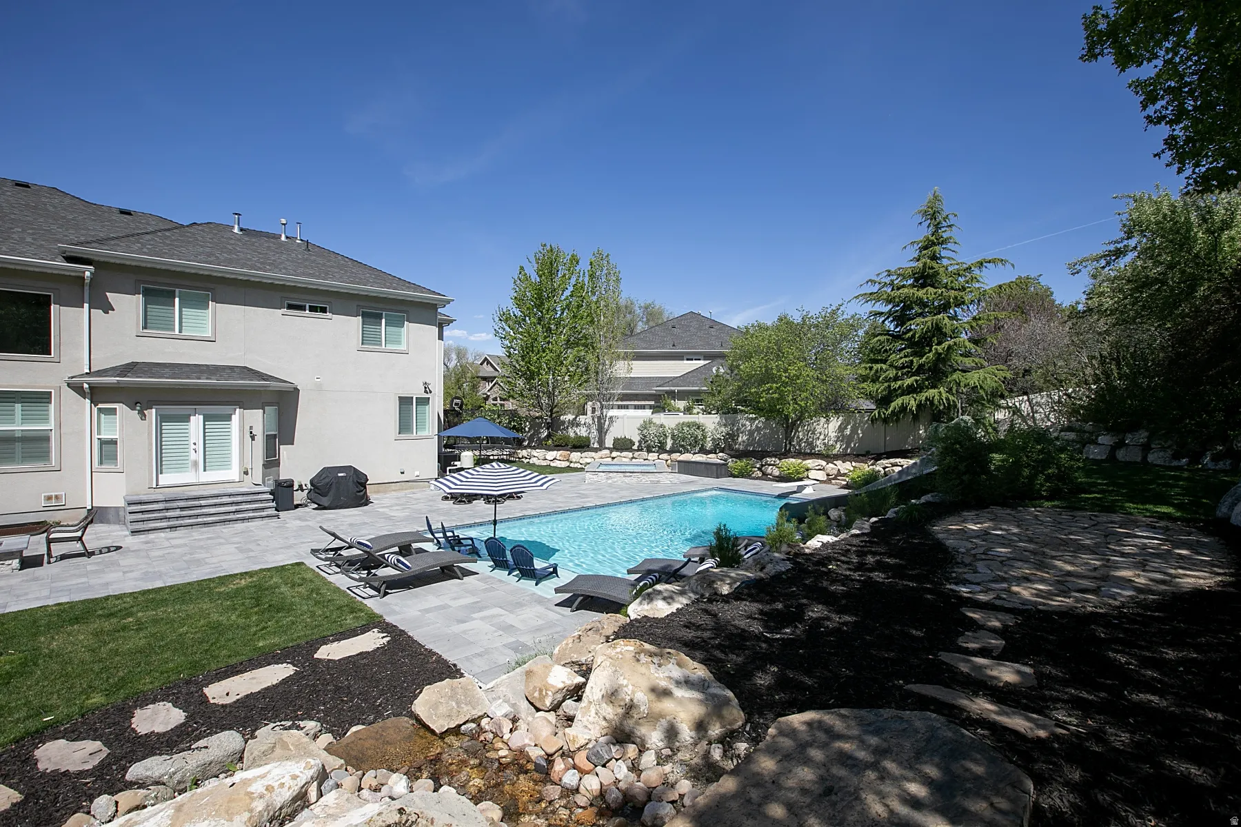 Expansive backyard featuring a large swimming pool with surrounding patio, a terraced landscape with mature trees, and a stone water feature