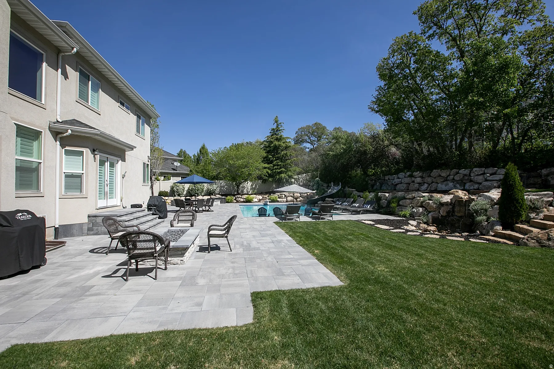 Expansive backyard featuring a large swimming pool, a tiered rock retaining wall, and a spacious paver patio with a built-in fire pit