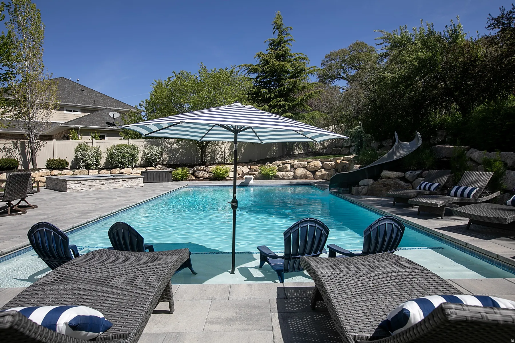 Expansive backyard featuring a rectangular swimming pool with a built-in slide, a perimeter of light-toned pavers, and a stone retaining wall