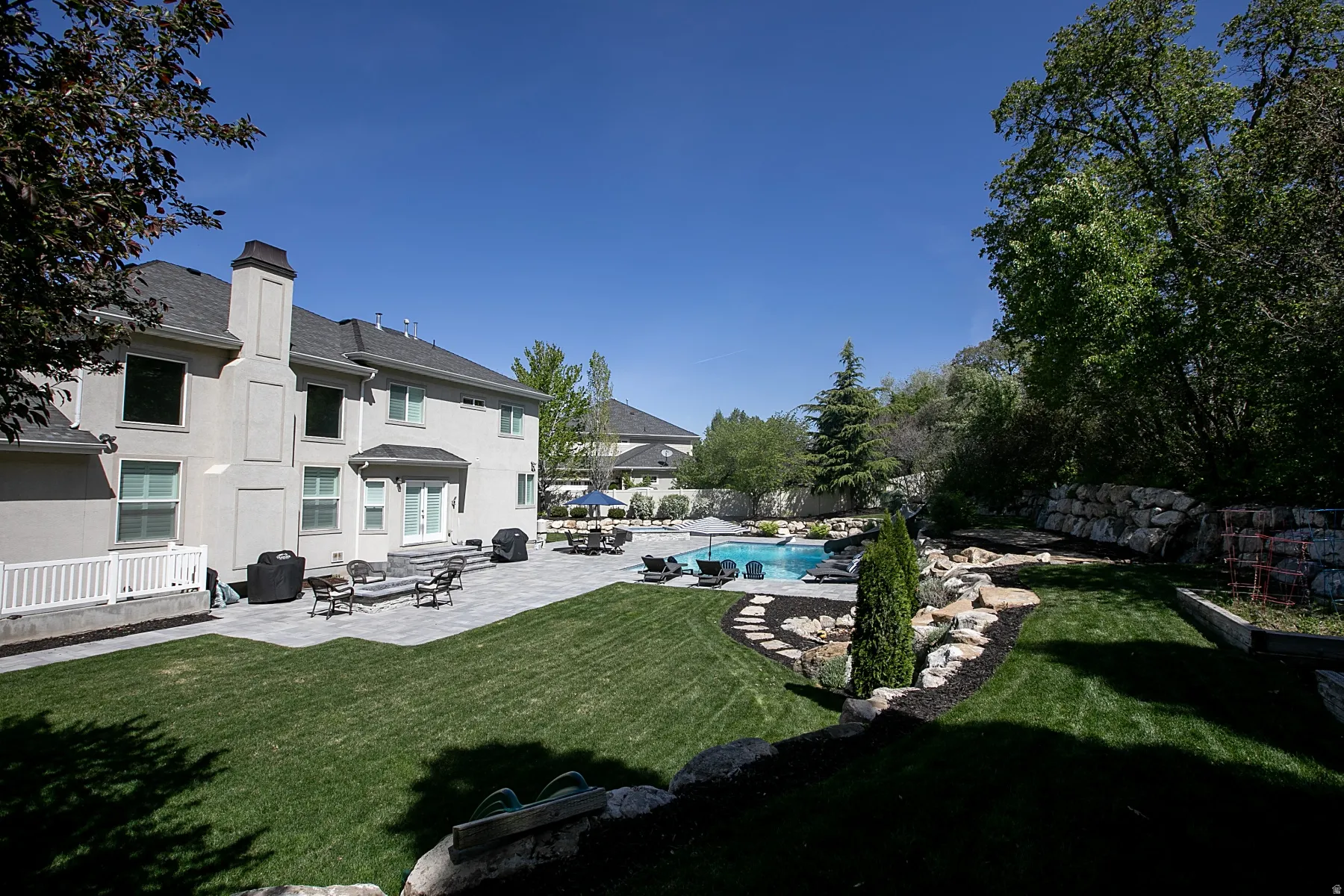Expansive exterior featuring a stucco facade, multiple windows, a large patio, an inground pool, and mature landscaping with rock features