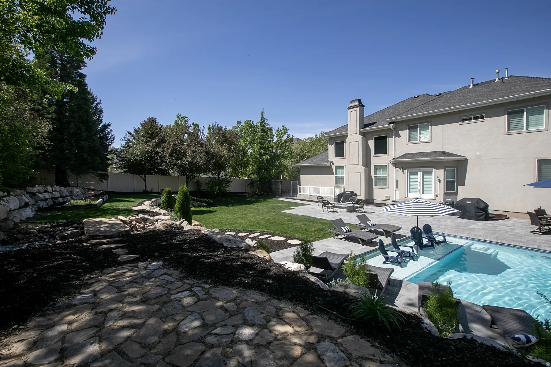 Spacious backyard featuring a rectangular swimming pool with integrated seating, extensive paver patio, stone retaining walls, and a manicured lawn