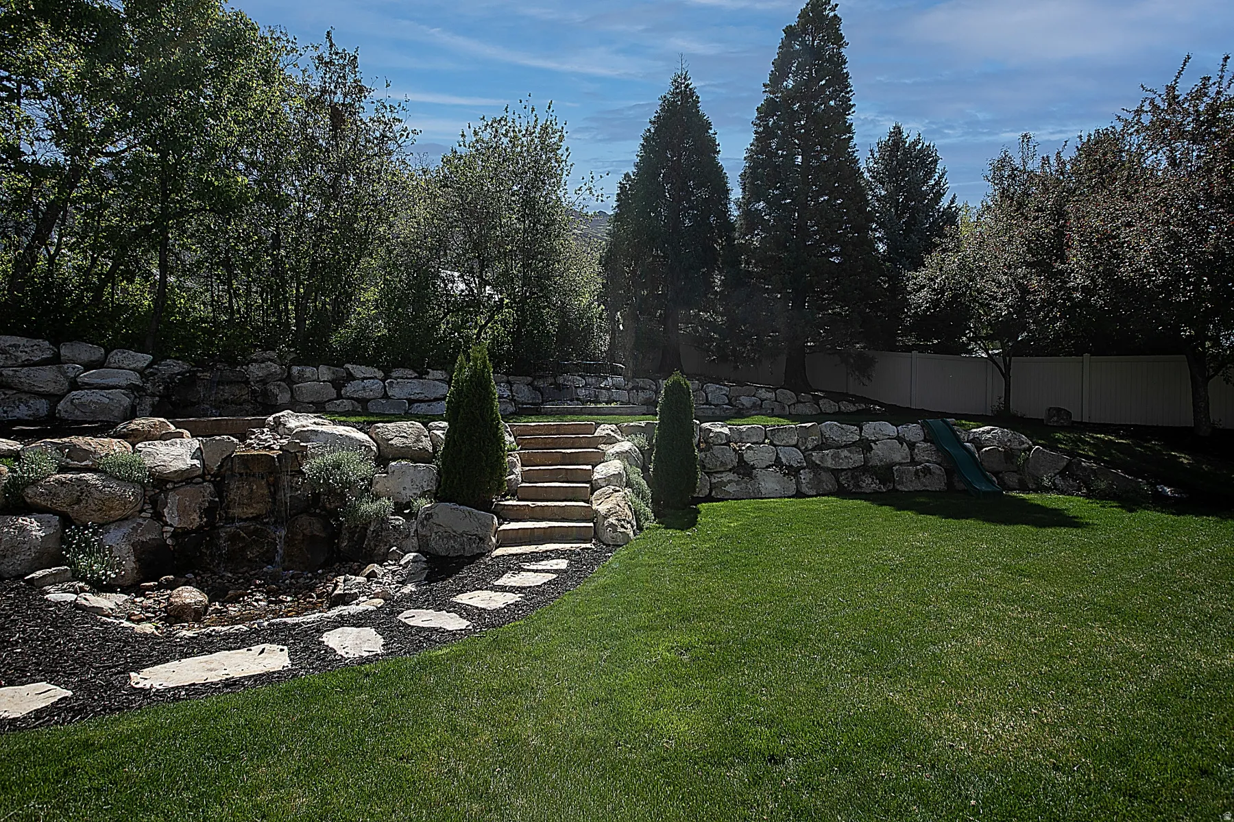 Expansive backyard featuring a lush lawn, multi-tiered rock retaining walls, a stone water feature, natural stone steps, and mature trees