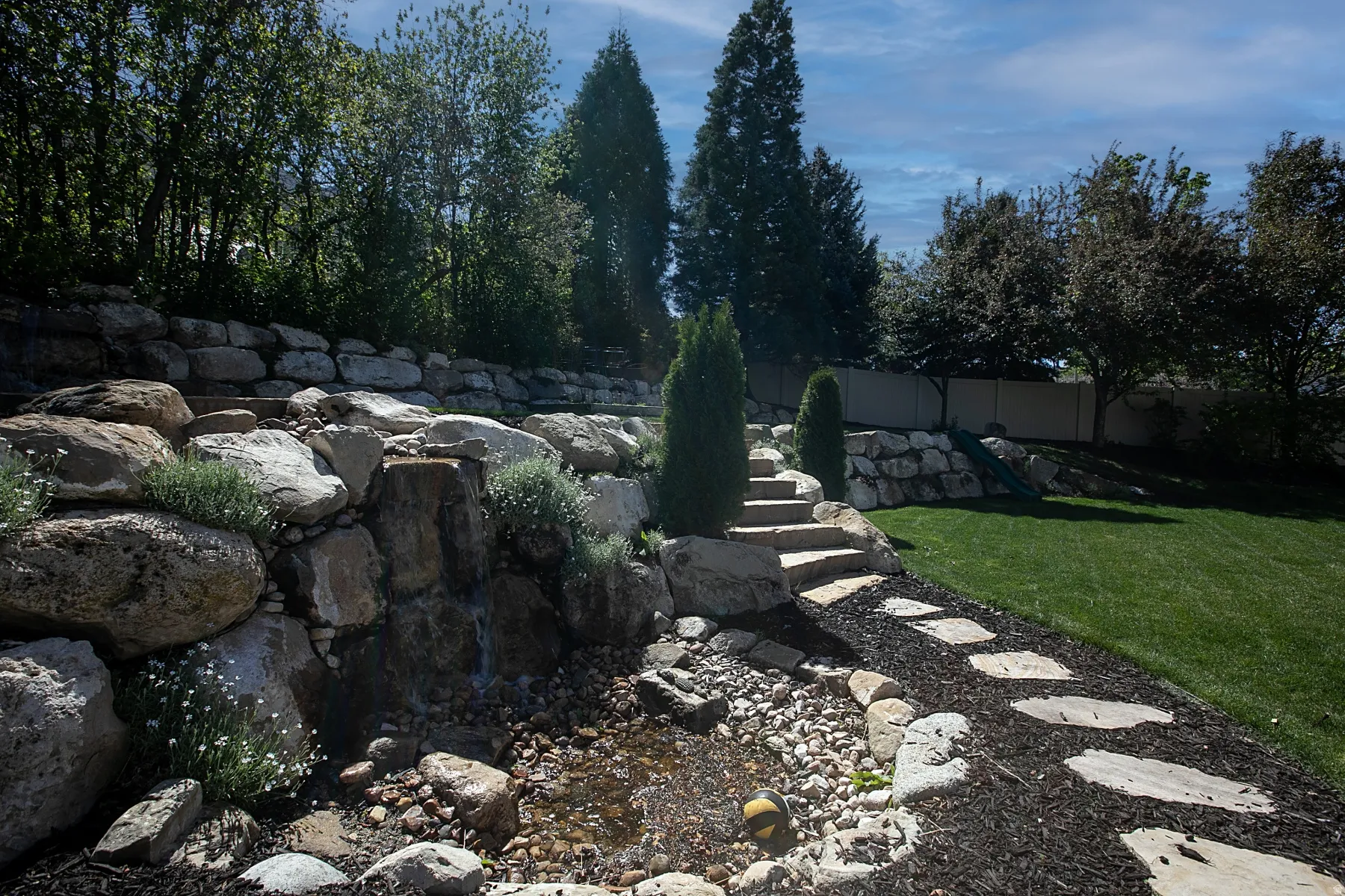 Tiered landscaping with a rock retaining wall, featuring a waterfall and a stepping stone pathway