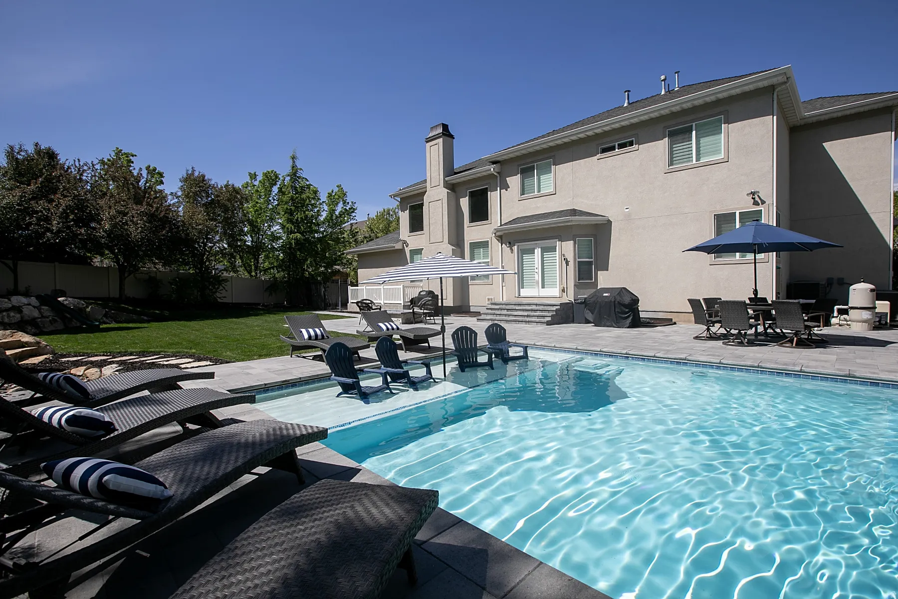 Expansive outdoor living area featuring a sparkling swimming pool with an integrated baja shelf and seating, a spacious paver patio, and mature landscaping