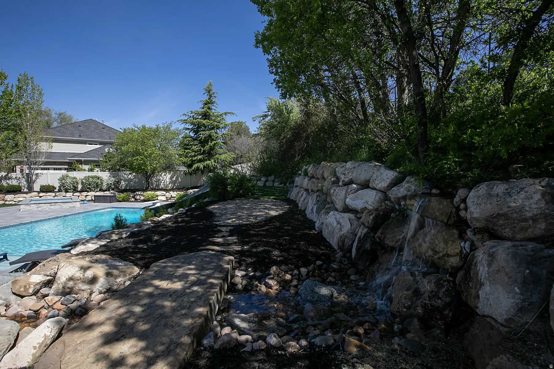 Outdoor oasis featuring a large swimming pool, rock retaining wall with cascading water feature, extensive landscaping, and mature trees