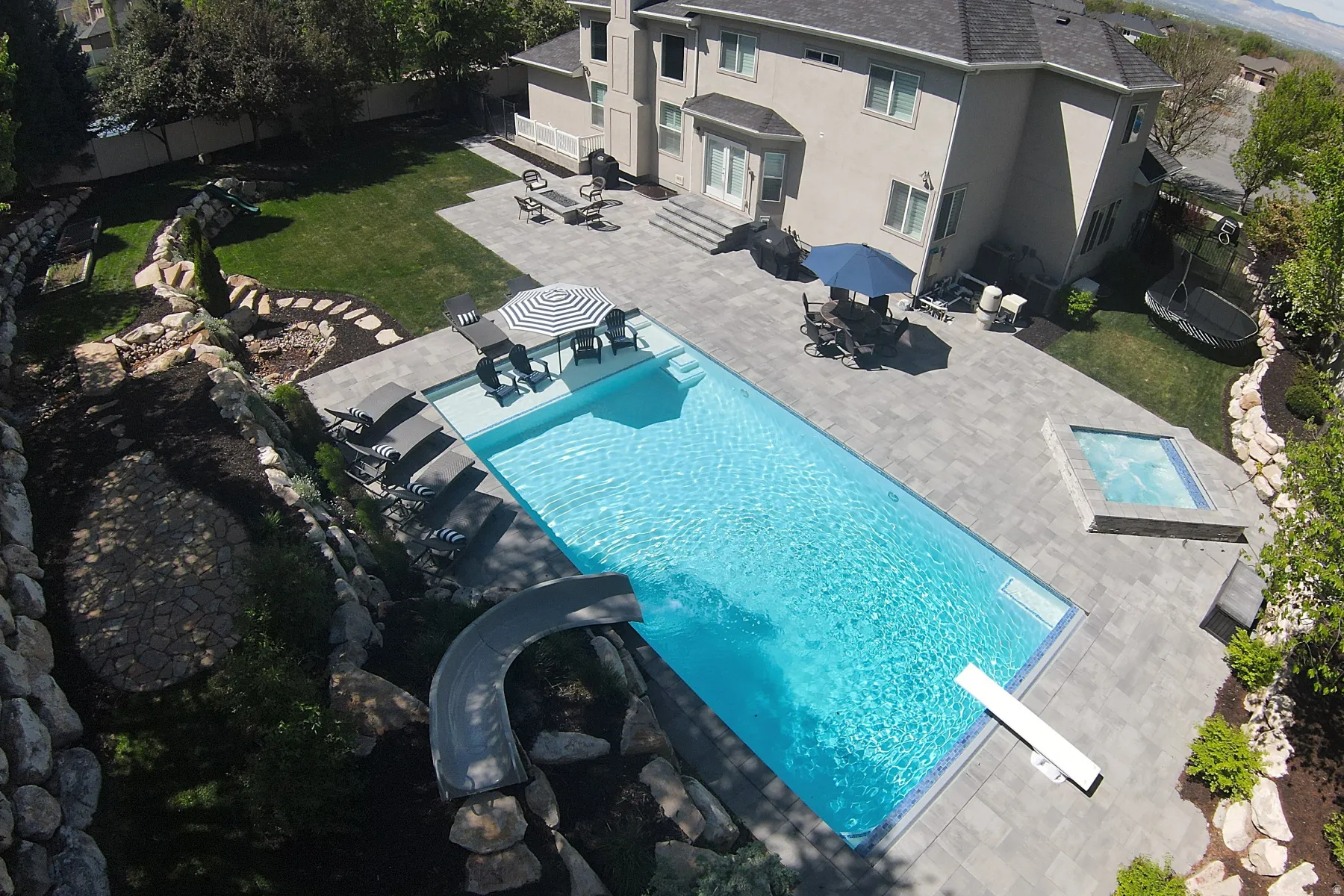 Backyard oasis featuring a large rectangular swimming pool with a diving board and slide, surrounded by extensive paver patio space