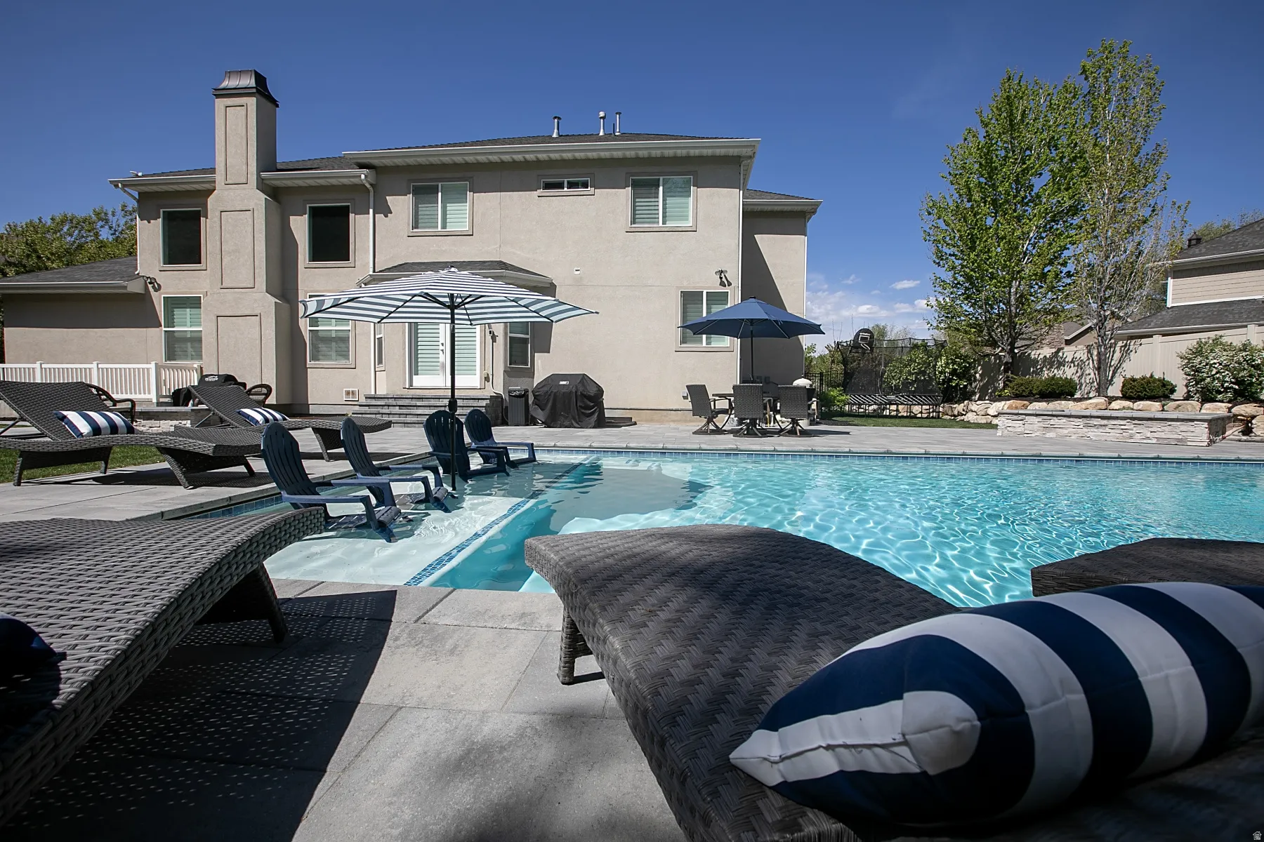 Backyard oasis featuring a large swimming pool with integrated tanning ledge, extensive stone patio, and stucco exterior with a prominent chimney stack