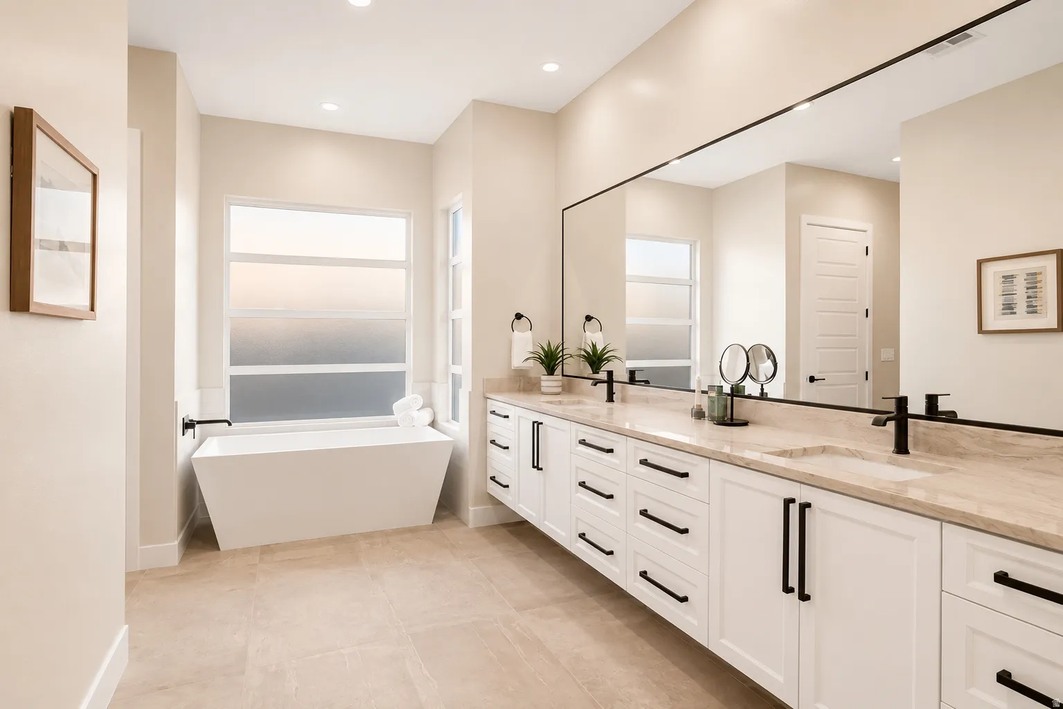 Contemporary bathroom featuring a freestanding soaking tub with a large frosted window, dual vanity with undermount sinks, stone countertops, extensive cabinetry with black hardware, and large format tile flooring