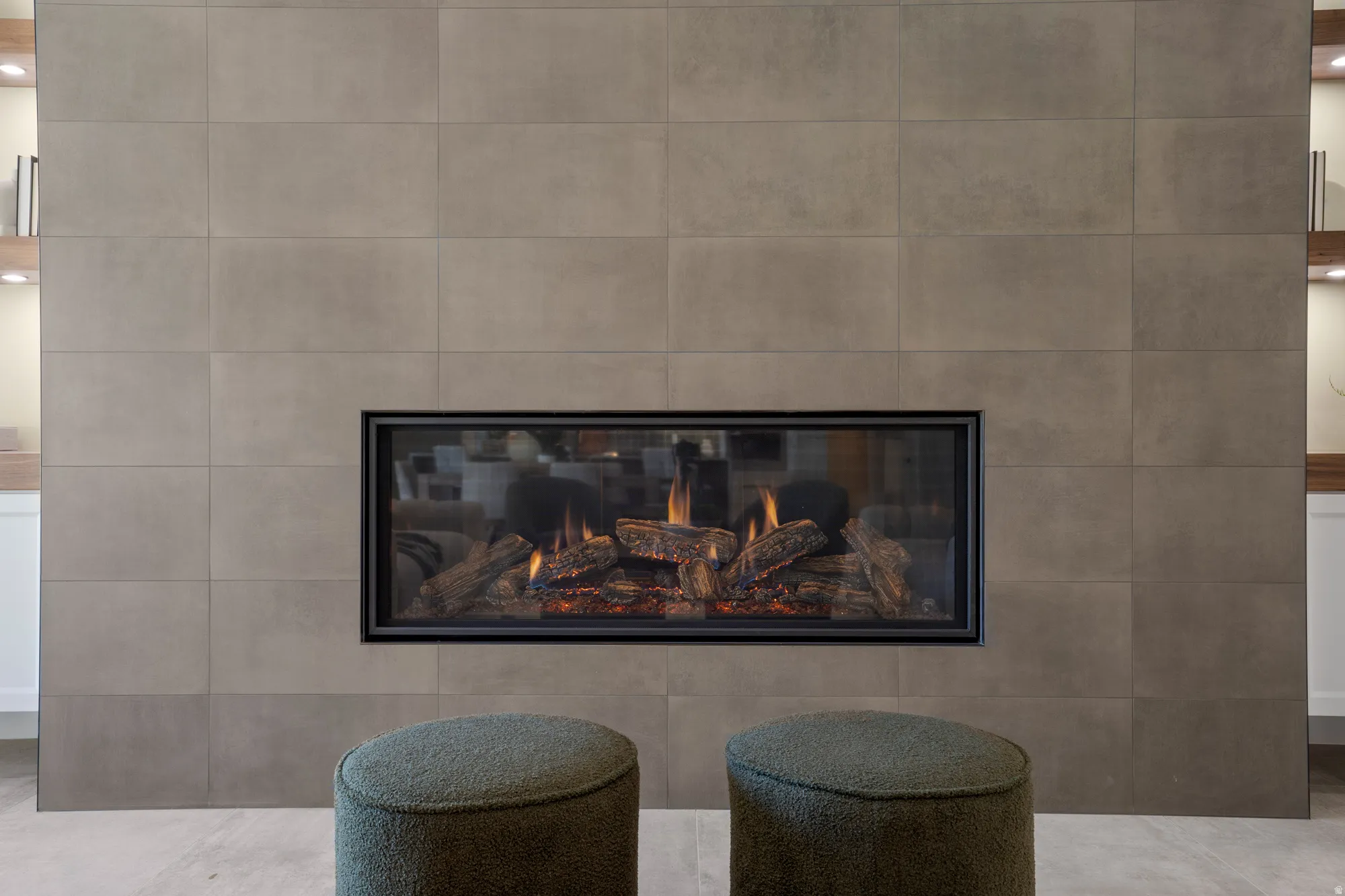 Modern fireplace featuring a wide glass-front insert with realistic log features, set within a large-format tile surround