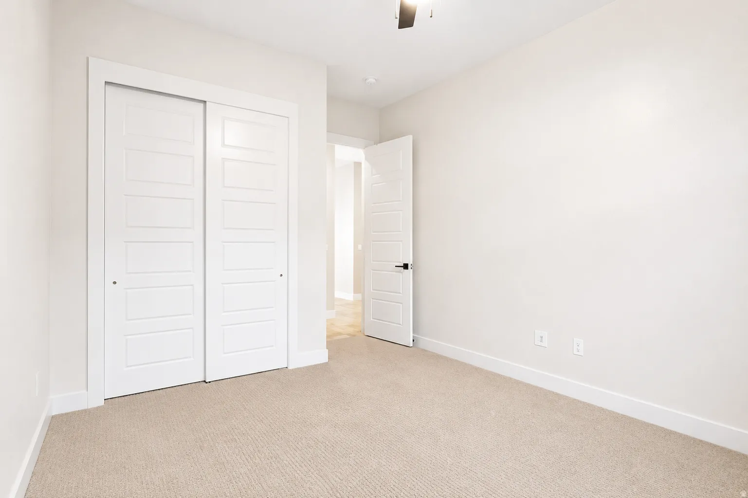 Neutral tone carpeting with white baseboards, a white paneled door with matte black hardware, and a ceiling fan with a dark finish