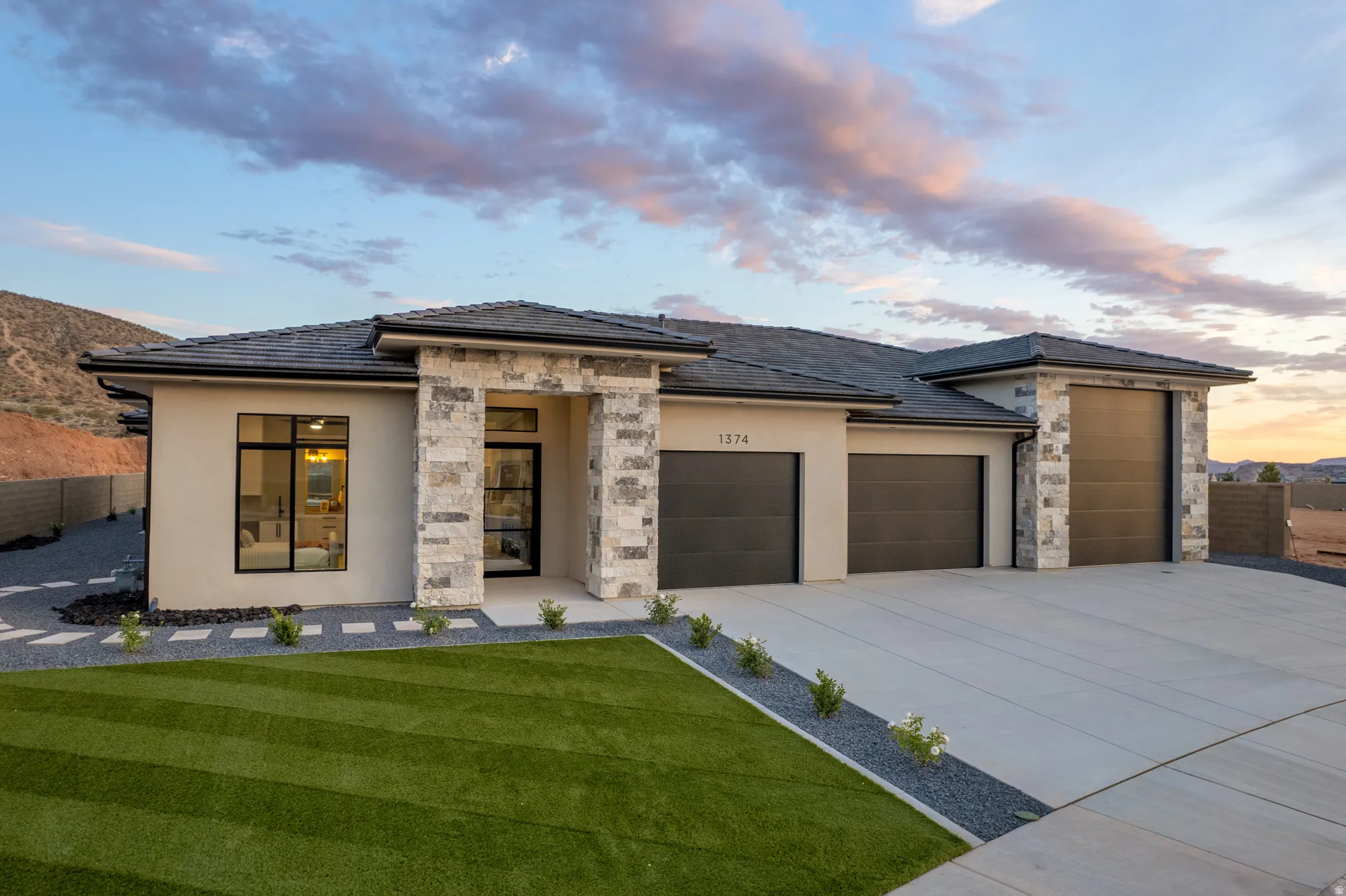 Contemporary stucco exterior with stone accents, featuring a multi-car garage, a flat concrete driveway, an artificial turf lawn, and a desert landscape backdrop