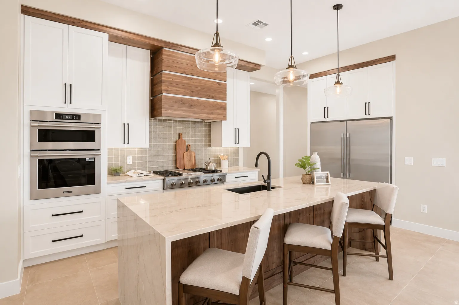 Gourmet kitchen featuring a large central island with a waterfall countertop, white cabinetry with contemporary hardware, stainless steel appliances including a built-in double oven, a gas range with a custom wood-finish hood, and tile flooring