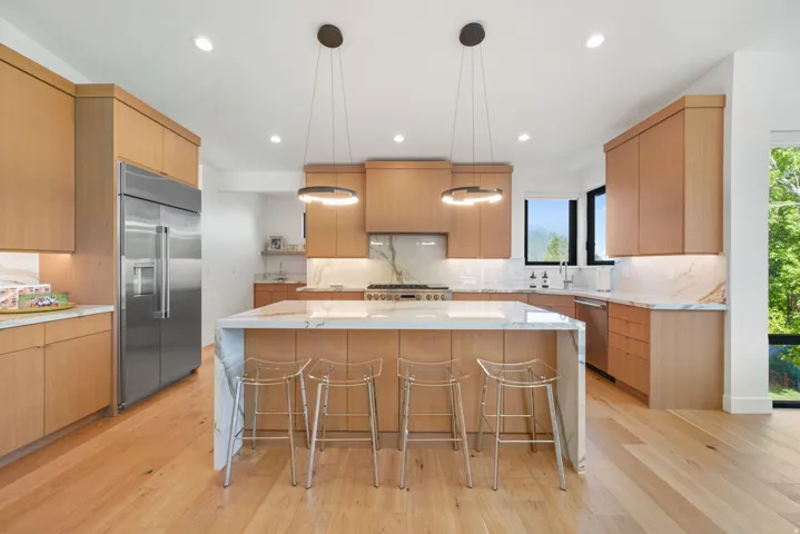 Gourmet kitchen featuring a large waterfall island with integrated seating, light wood-finish cabinetry, stainless steel appliances, and a full-height marble-style backsplash