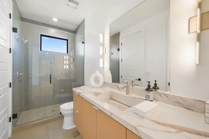 Modern bathroom featuring a spacious glass-enclosed shower with a rectangular window, a light wood-finish vanity with ample storage, a stone-finish countertop, a rectangular vessel sink, and a large wall-mounted mirror with integrated lighting