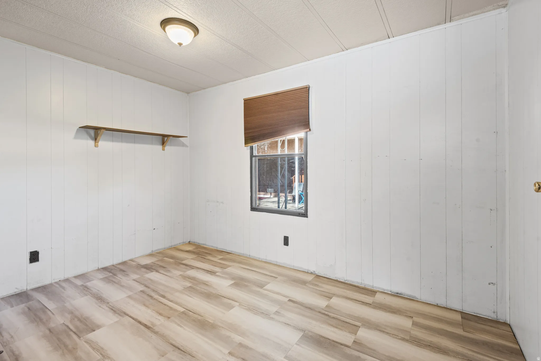 Room featuring wood-finish flooring, white vertical paneled walls, and a recessed window with a brown blind