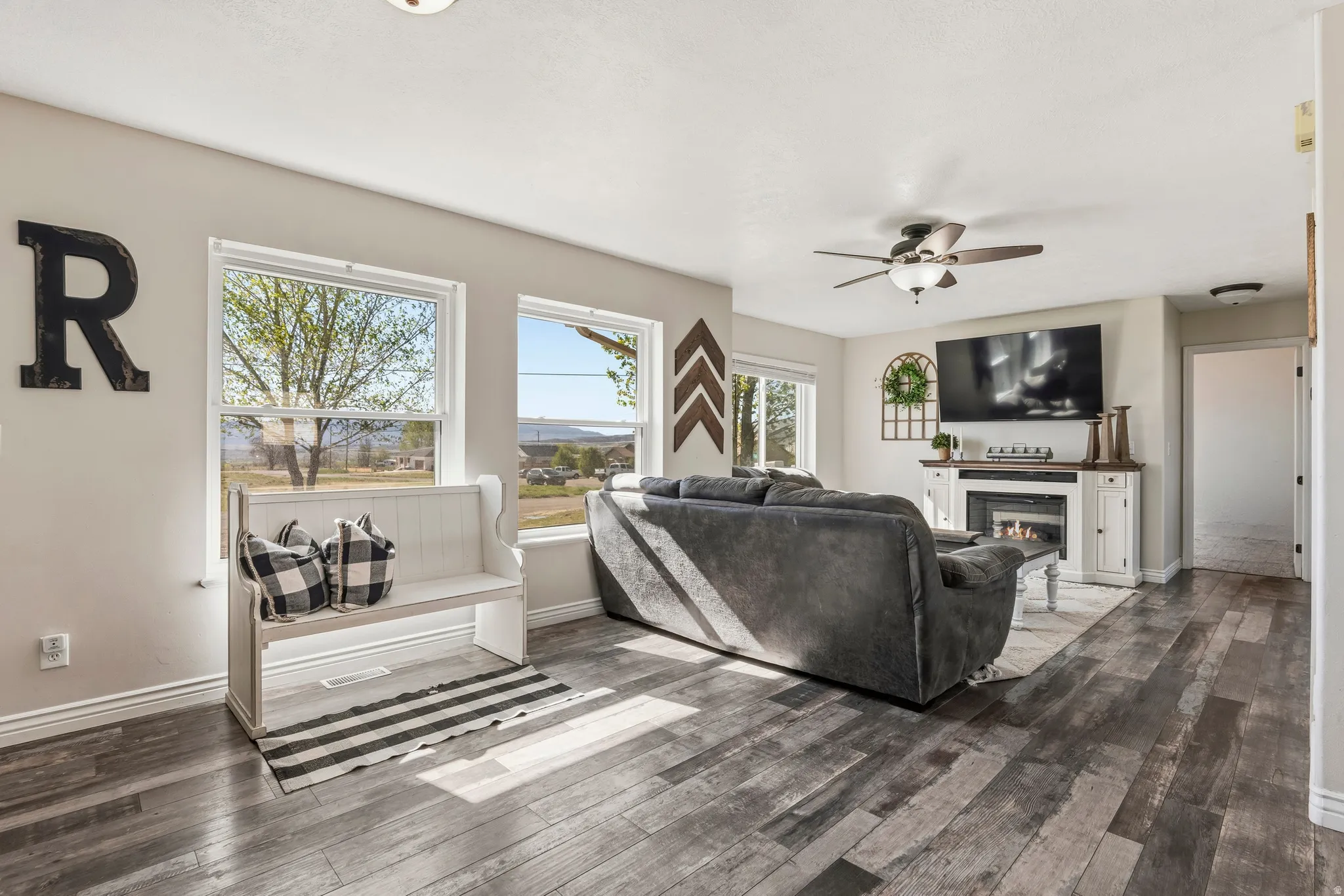 Spacious living area featuring wood-finish flooring, multiple windows, a ceiling fan, and a built-in electric fireplace with mantel and cabinetry