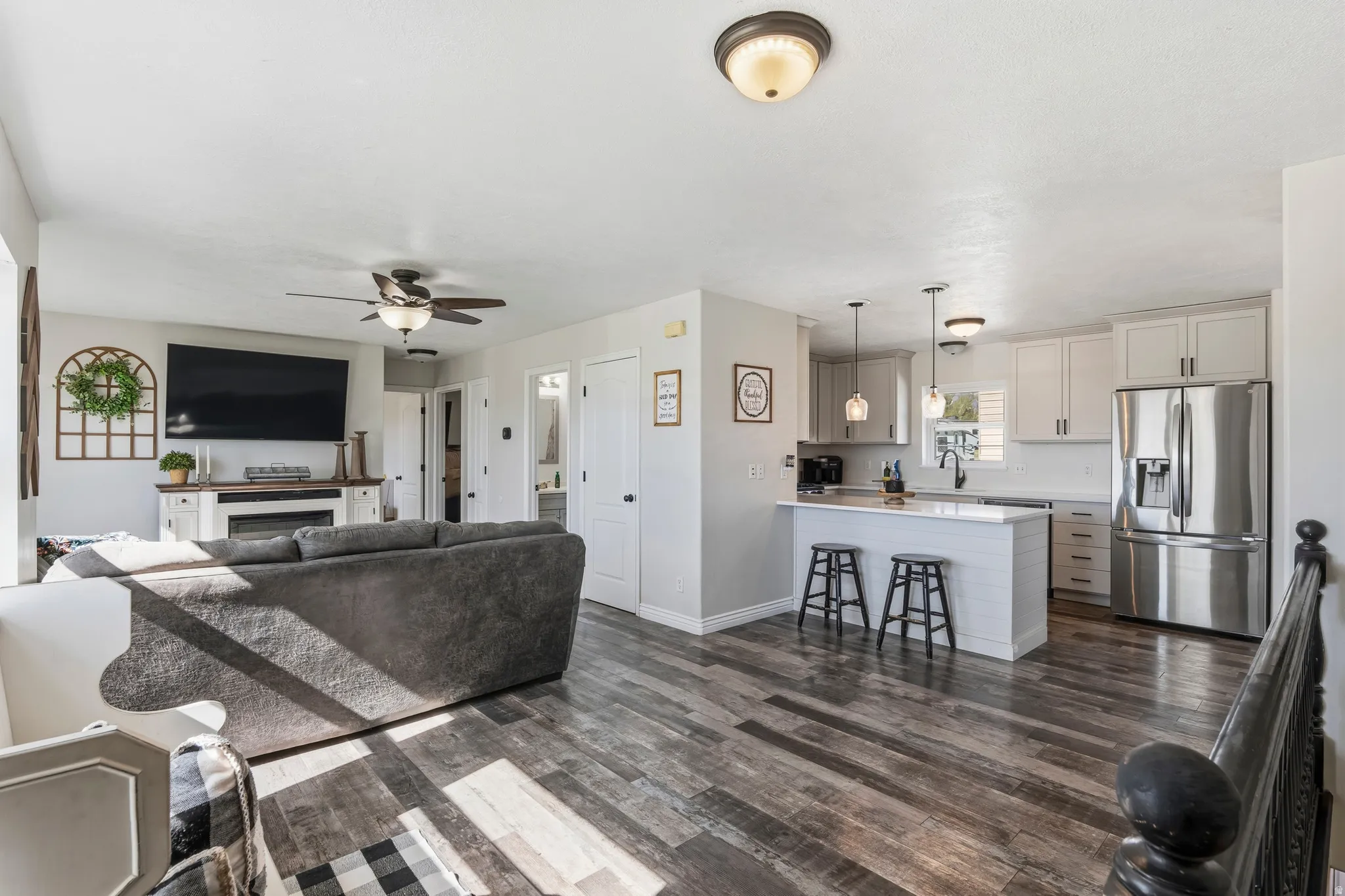 Open-concept living space with wood-finish flooring, a contemporary kitchen island with seating, pendant lighting, stainless steel appliances, and a built-in electric fireplace with mantel