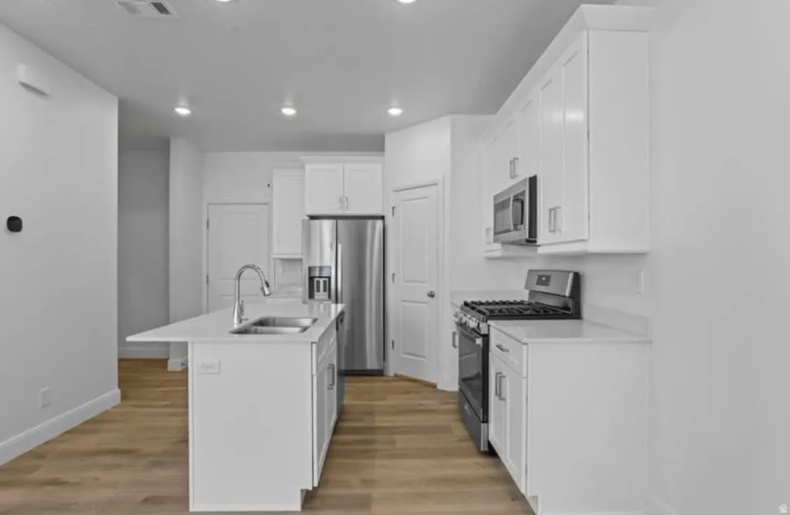 Kitchen featuring an island with an undermount sink, white shaker cabinetry, stainless steel appliances, and wood-finish flooring