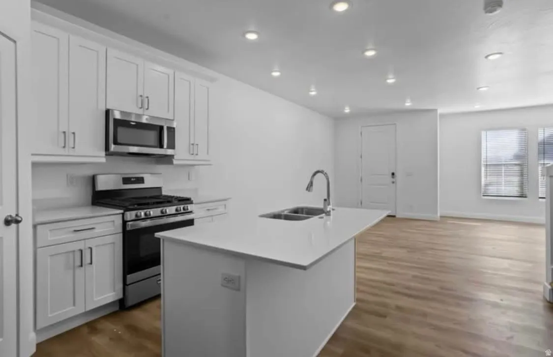 Modern kitchen featuring white shaker cabinetry, stainless steel appliances, a center island with a double basin sink, recessed lighting, and wood-finish flooring