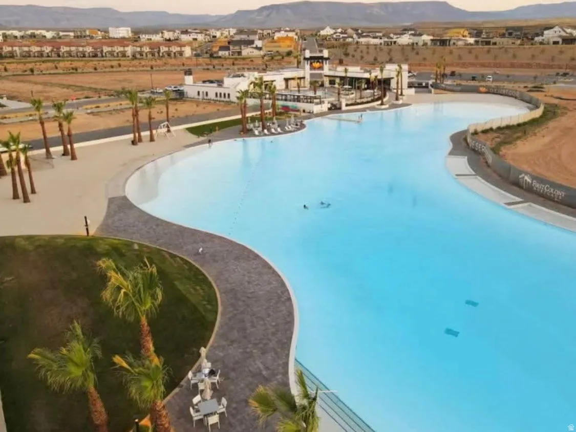 Expansive lagoon-style swimming pool featuring a white sand beach entry, surrounding paved walkways, established palm trees, and a distant community building with a multi-level deck