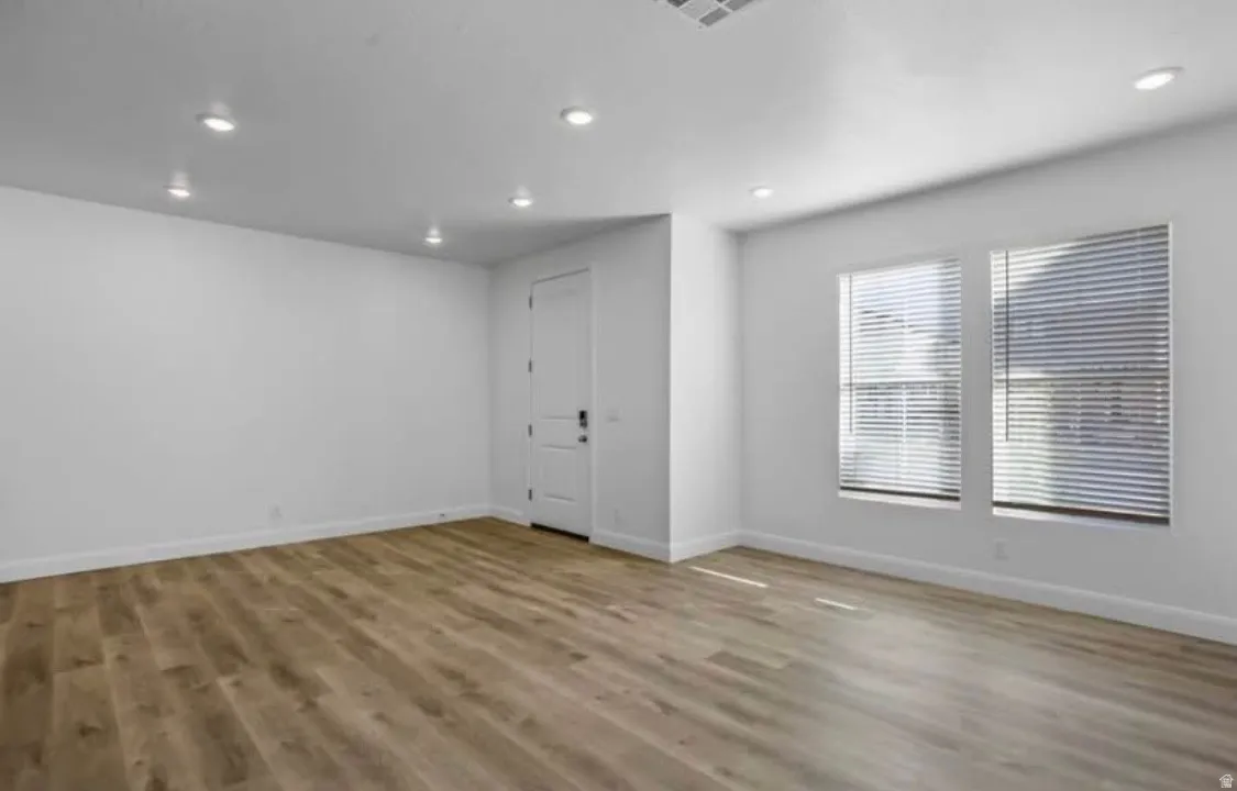 Spacious room featuring wood-finish flooring, recessed ceiling lighting, two windows with blinds, and white walls