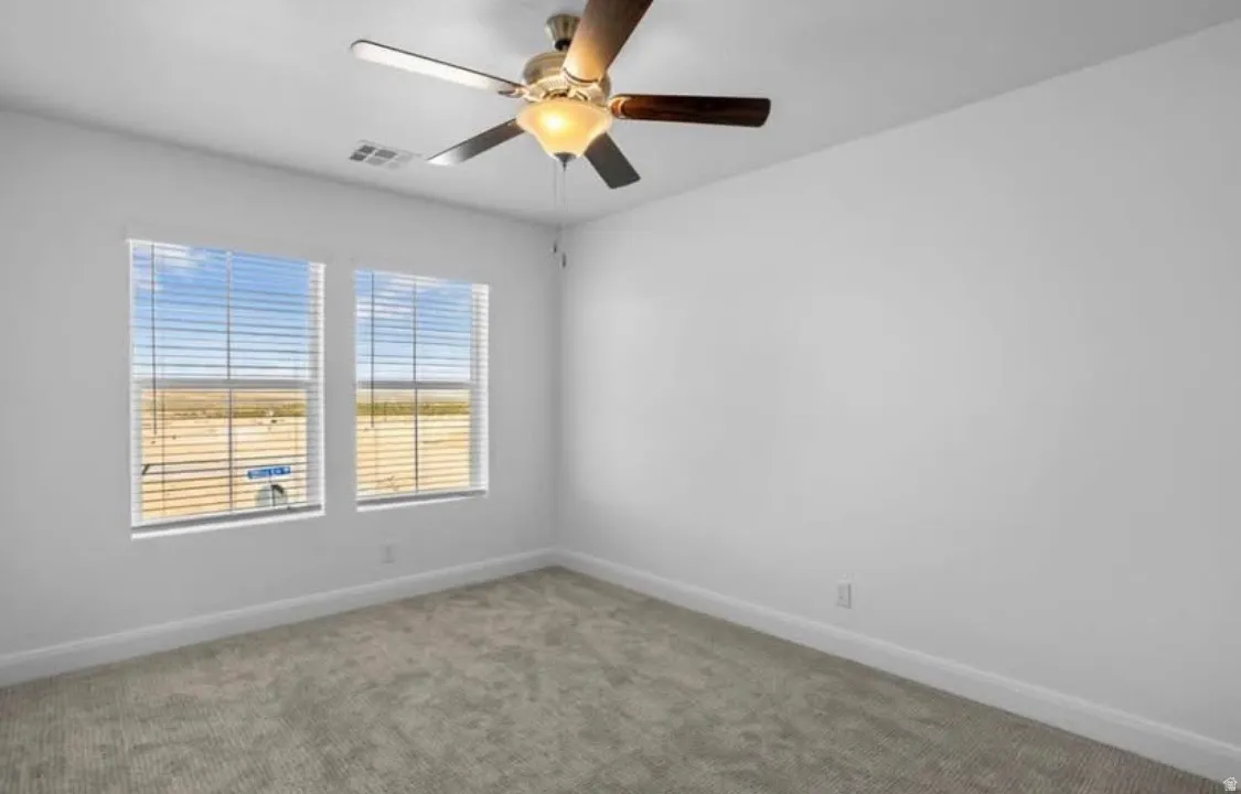 Spacious room featuring dual windows with blinds, a ceiling fan with integrated lighting, plush grey carpeting, and clean white walls