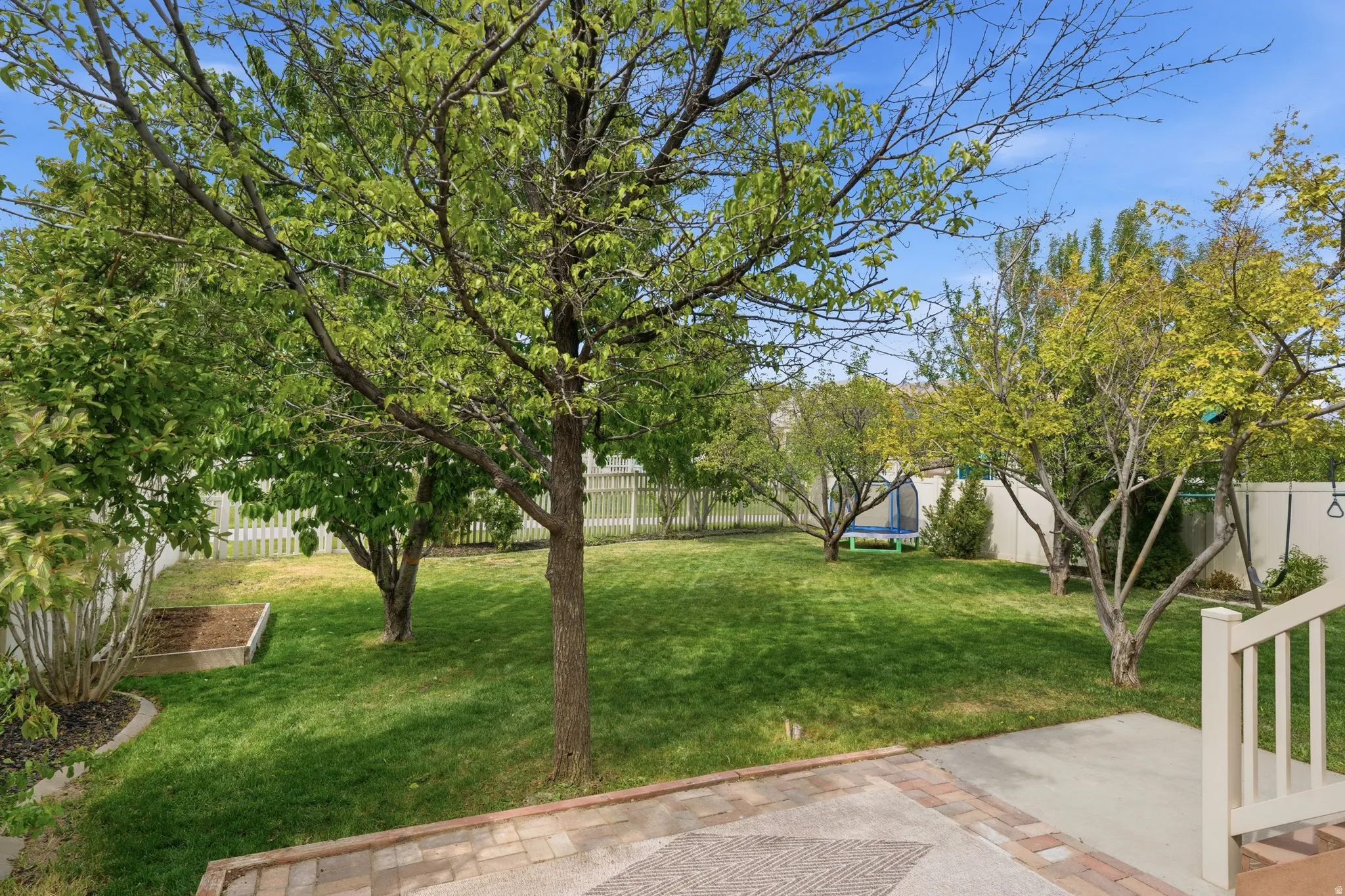 Expansive backyard featuring a green lawn, mature trees, a white picket fence, a raised garden bed, and a paved patio area
